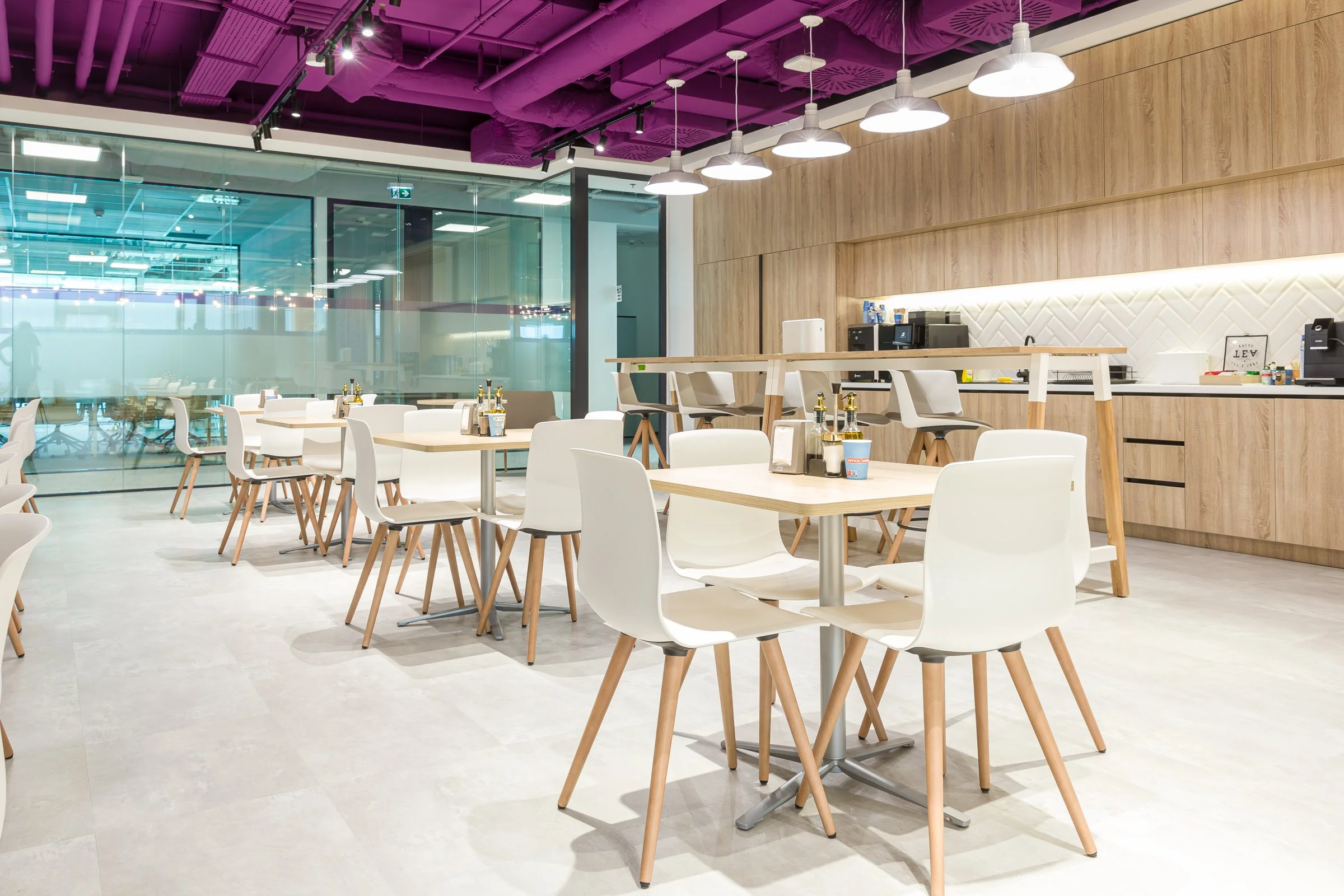 Empty modern cafe with white chairs, wooden tables, and a bar area with coffee machines, illuminated by hanging white lights, with a glass wall and purple ceiling accents.