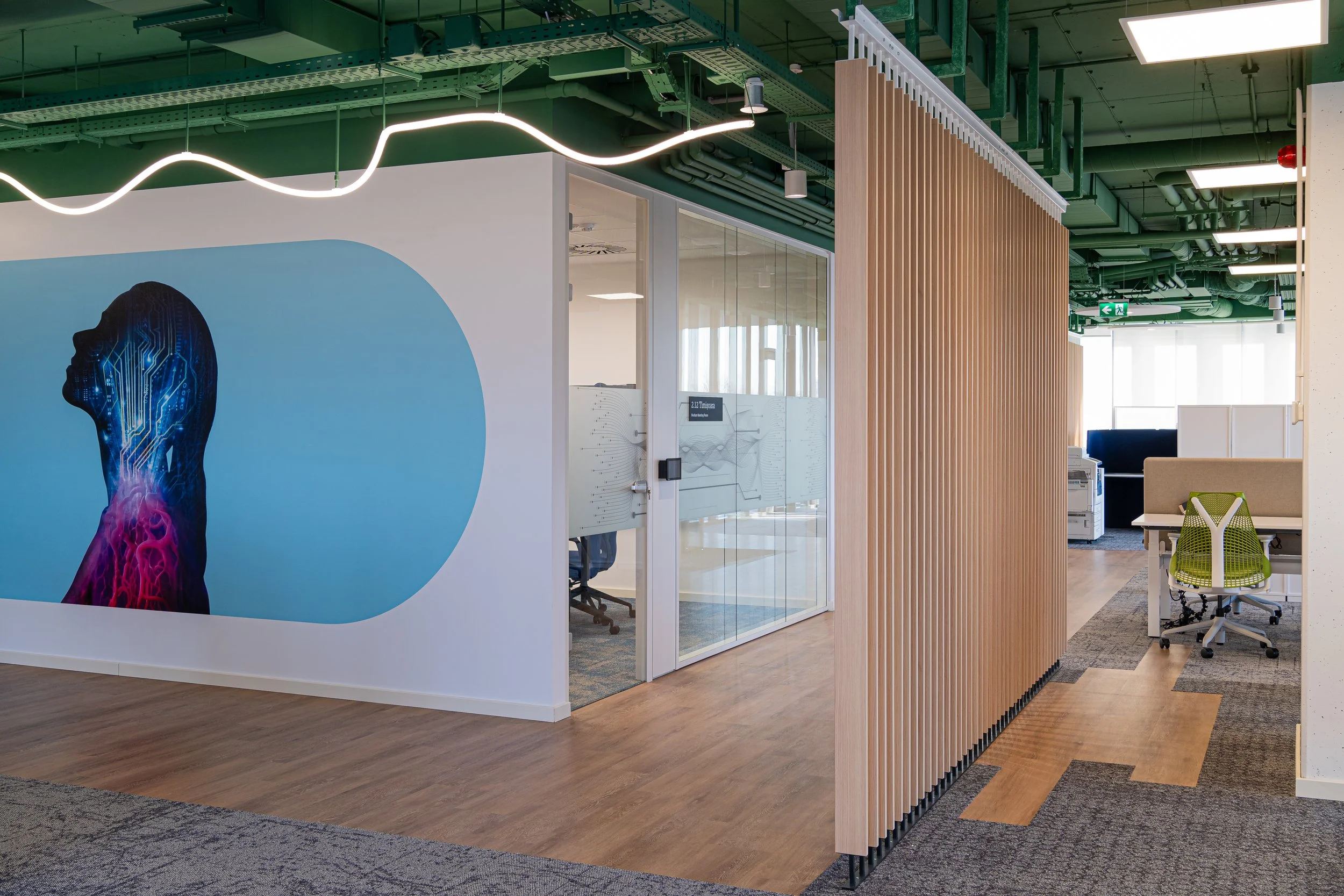 Modern office space with a wall mural of a digital human head, glass-enclosed rooms, wooden slat divider, and workstations with green chairs.