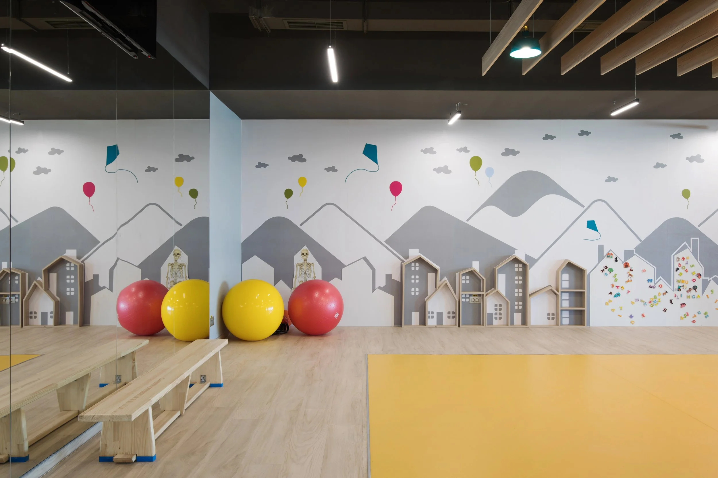 A room with a mural of mountains, clouds, and balloons on the wall. There are gym balls, skeleton mannequins, small house-shaped shelves, and a yellow padded area on the floor.