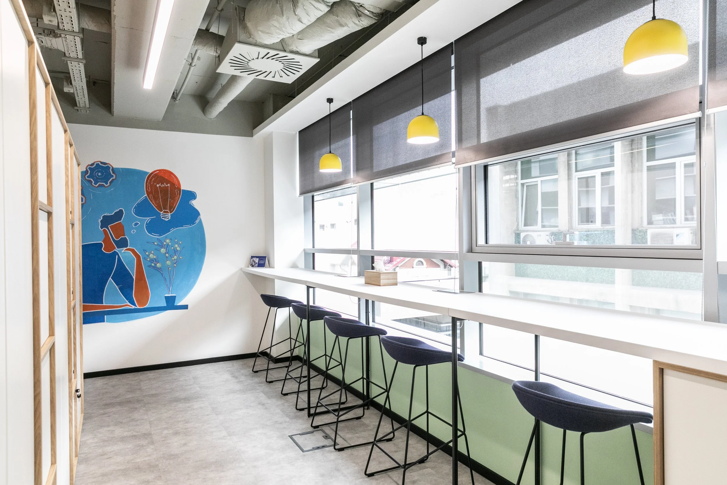 Modern coworking space with high bar stools along a windowed wall, gray blinds, yellow pendant lights, and a colorful mural on the white wall showing a person thinking with a lightbulb above their head.