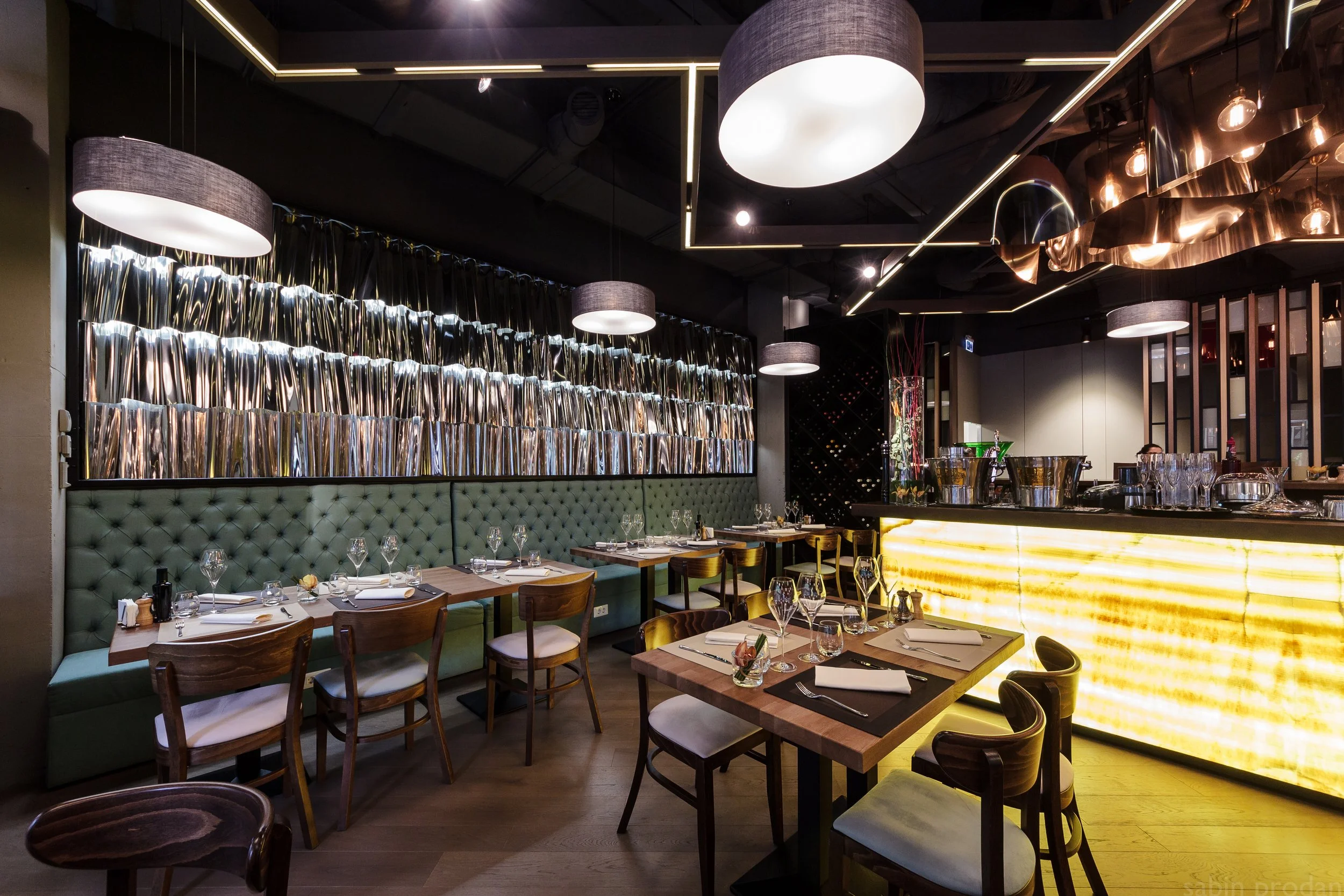 Elegant restaurant interior with wooden tables, green tufted banquette, wine glassware, modern pendant lights, and a brightly illuminated bar counter.