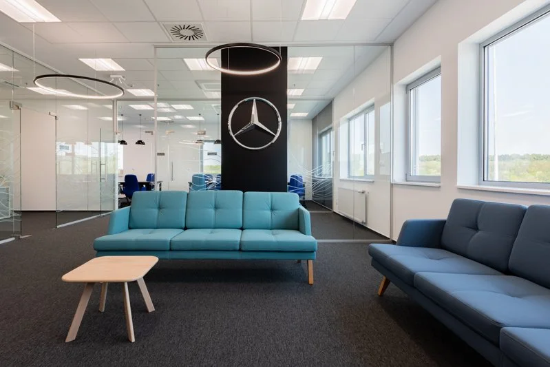 Modern office lounge area with two blue sofas, a small wooden coffee table, and large windows, featuring a Mercedes-Benz logo clock on a black column.