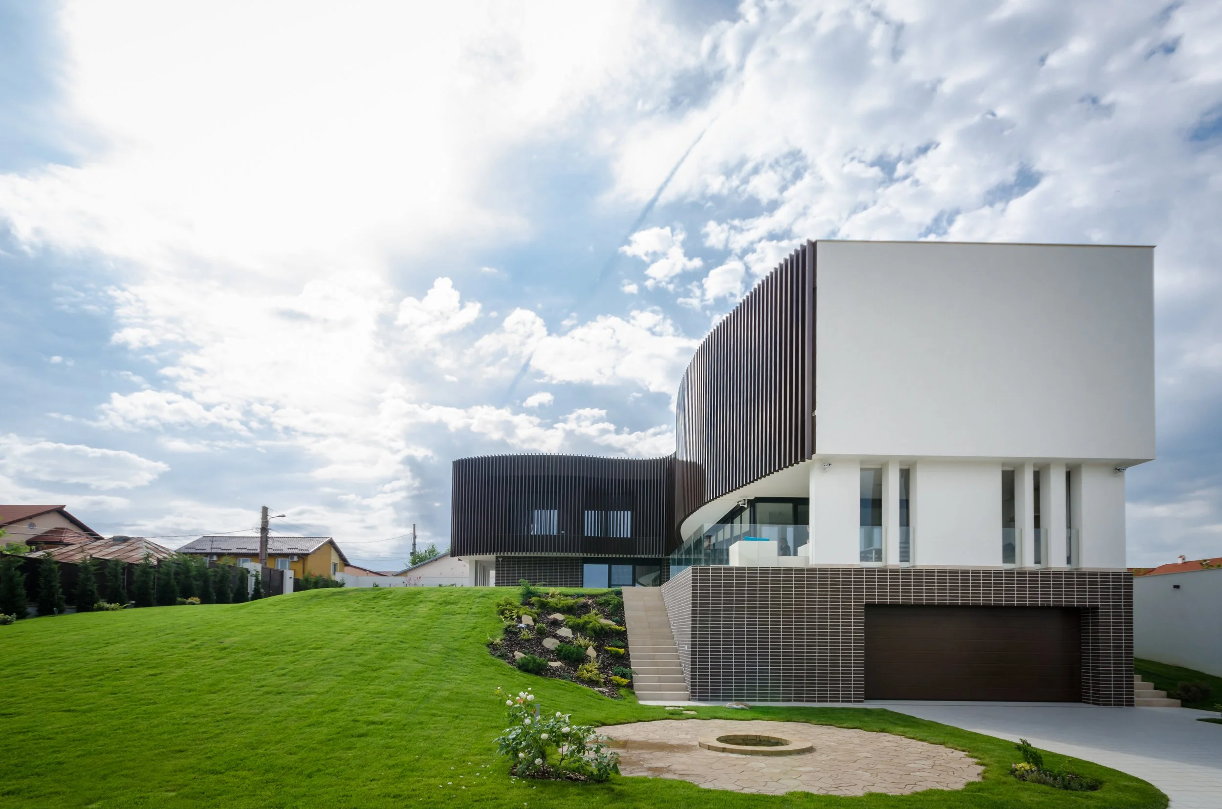 Modern house with curved black and white exterior walls, large windows, and a garage, set on a green lawn under a cloudy sky.