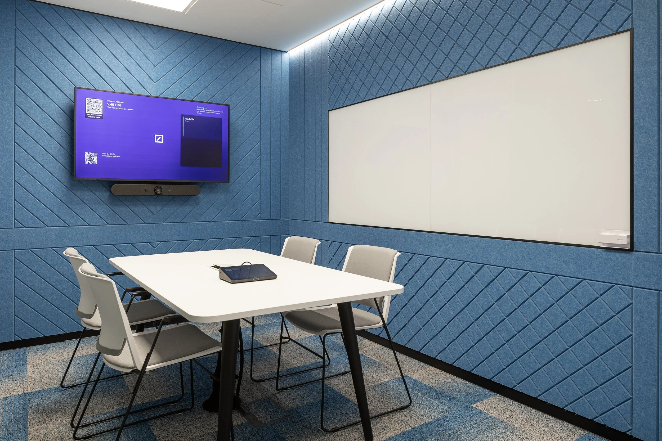 A modern meeting room with a white table, five grey chairs, a large whiteboard, and a mounted flat-screen TV on a textured blue wall.