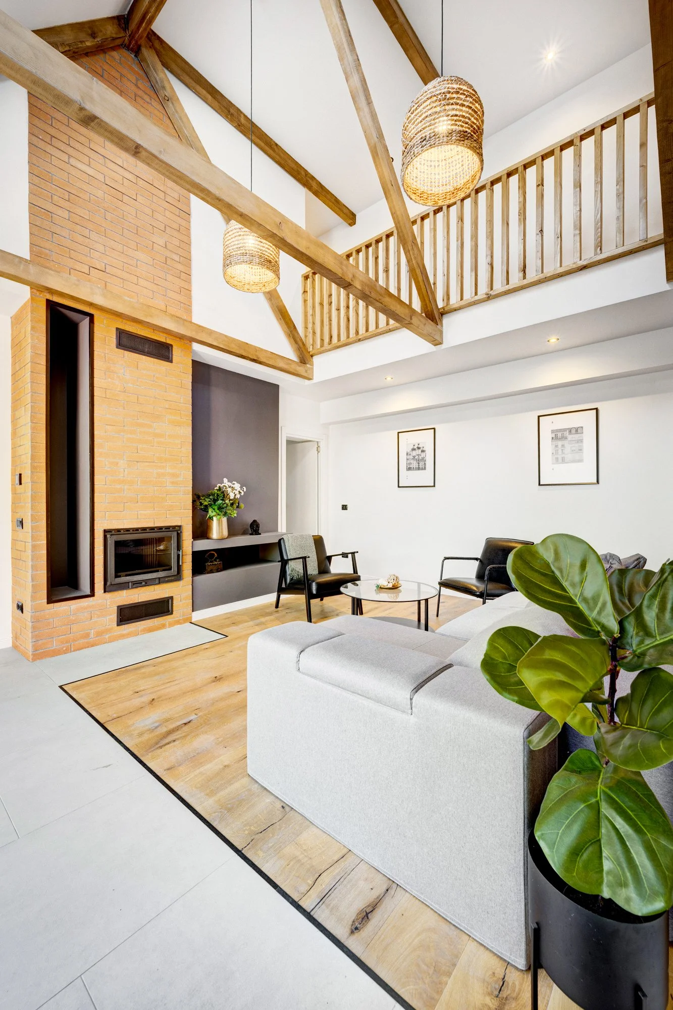 Modern living room with white walls, a brick fireplace, wooden beams, hanging wicker lights, a gray and white sofa, black armchairs, a round glass coffee table, framed artwork on the wall, and green leafy plants.