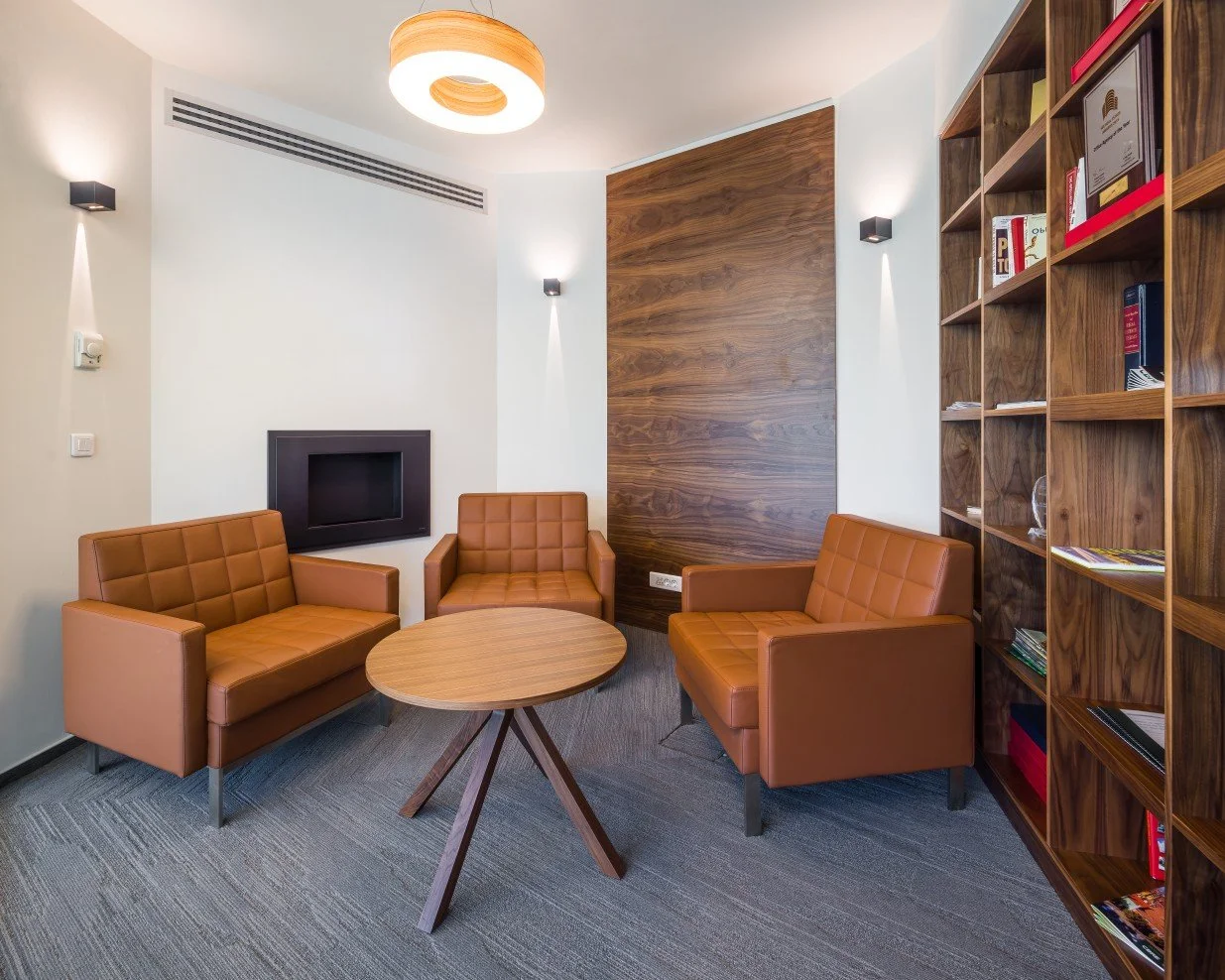 A small waiting area with three tan leather armchairs around a round wooden table in a modern room with white walls, a wooden accent wall, and built-in bookshelf filled with books and magazines.