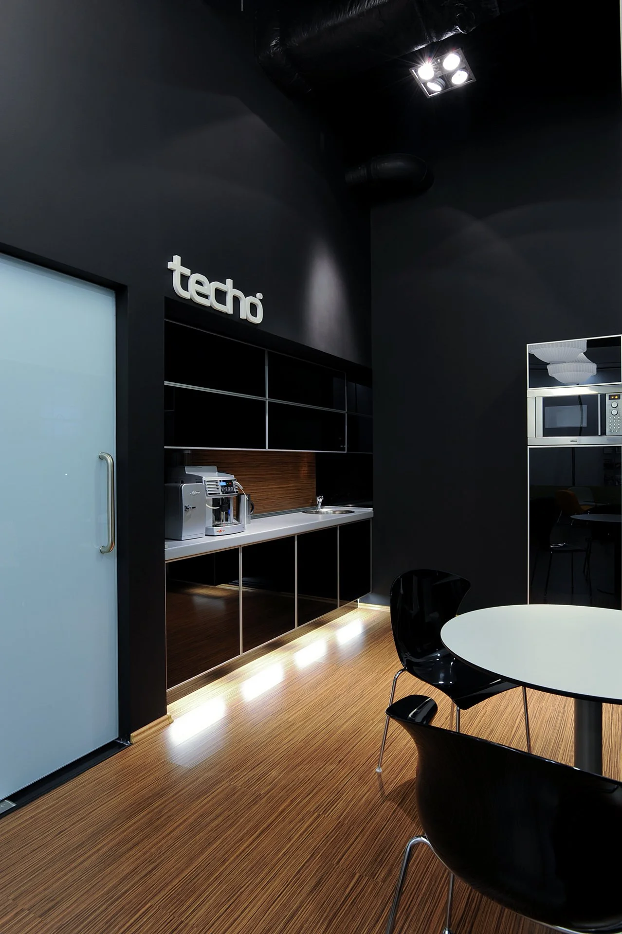 Modern office kitchenette with black walls, wooden flooring, a white round table, black chairs, a frosted glass door, and kitchen appliances including a coffee machine and microwave, with the sign 'techo' on the wall.