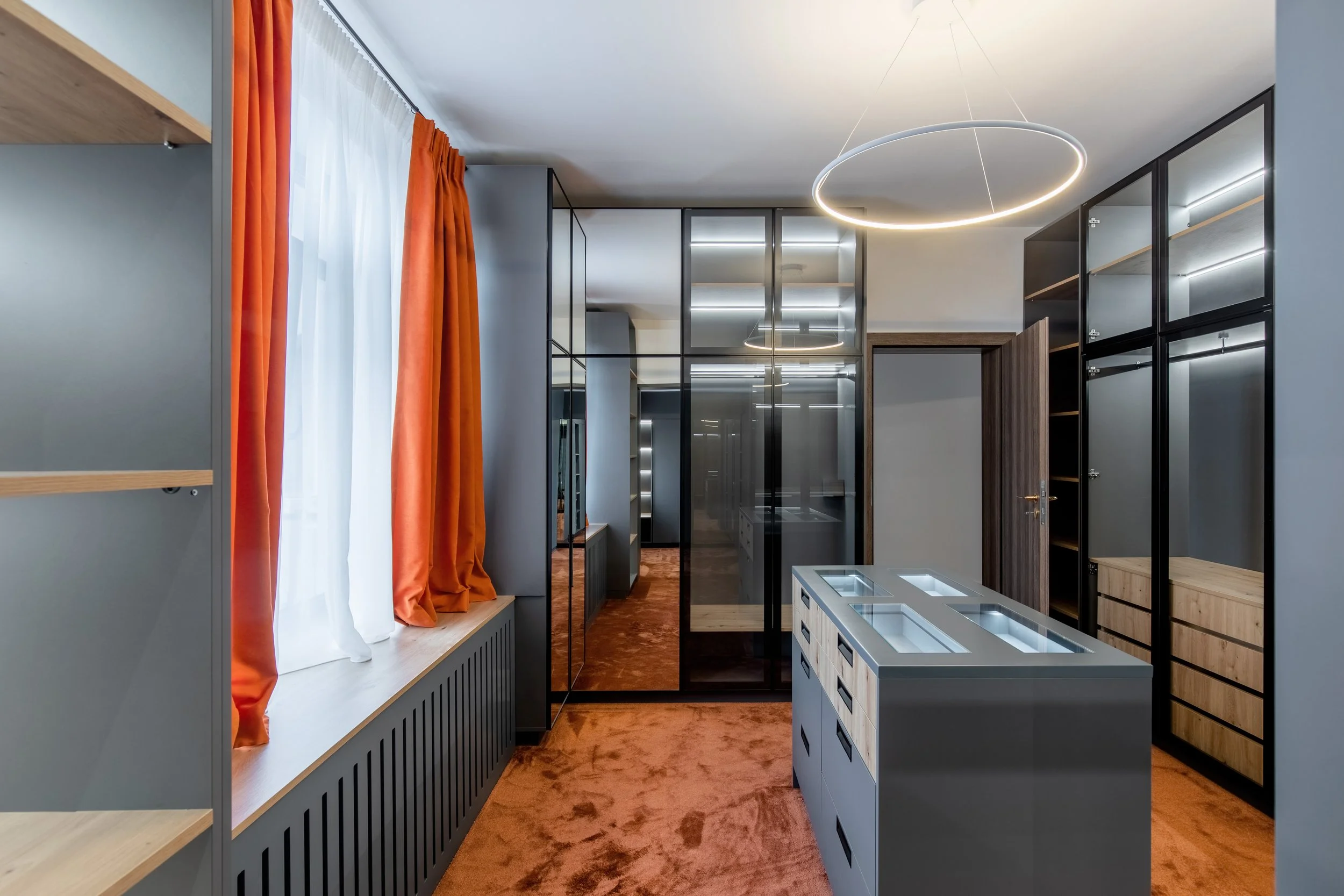 Modern kitchen with gray cabinetry, black glass cabinets, a central gray island with glass display sections, orange curtains over a window, and circular ceiling light fixtures.