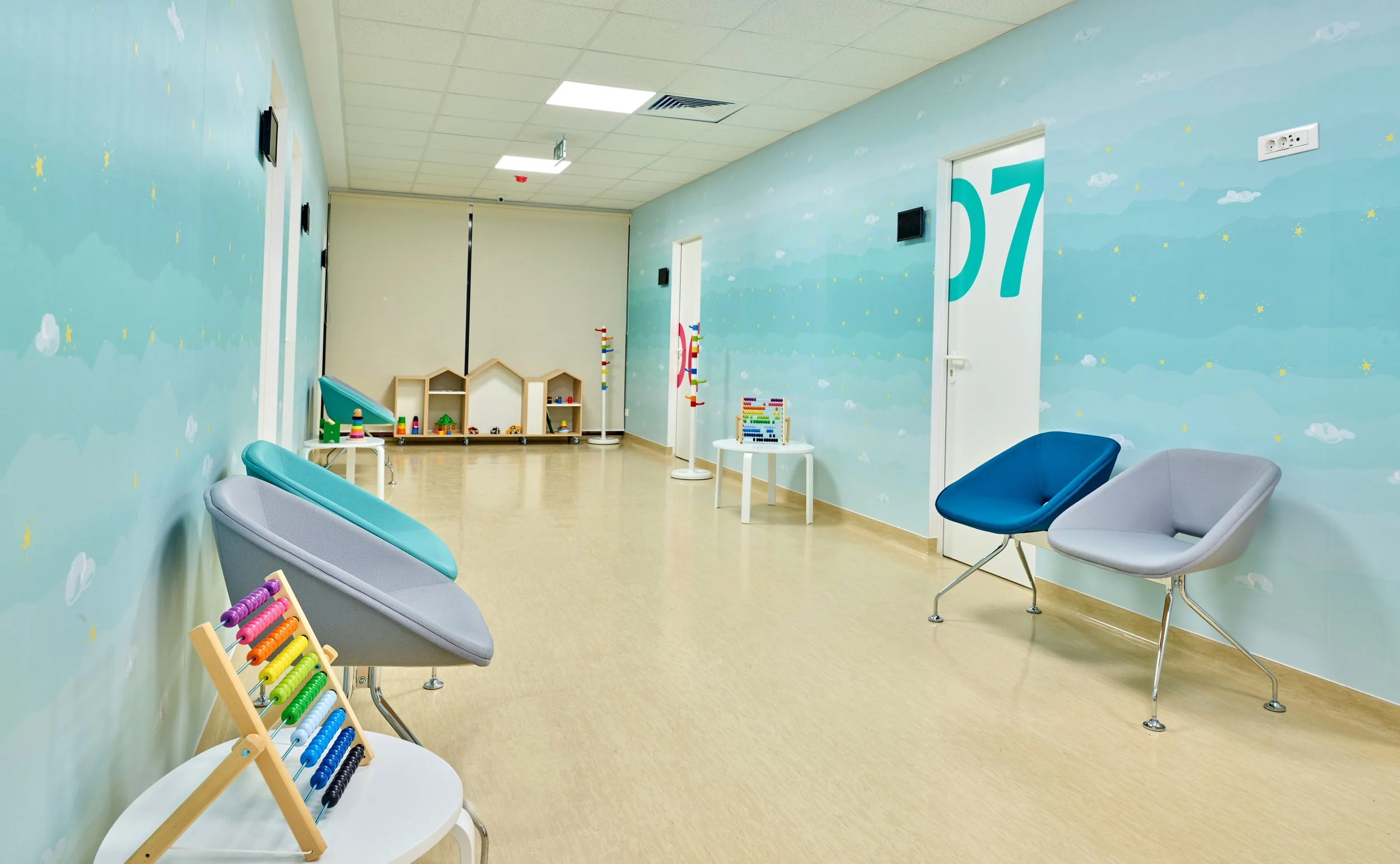 Empty waiting area with pastel-colored chairs, a toy abacus, and soft blue clouds and stars painted on the walls, in a children's medical or dental office.