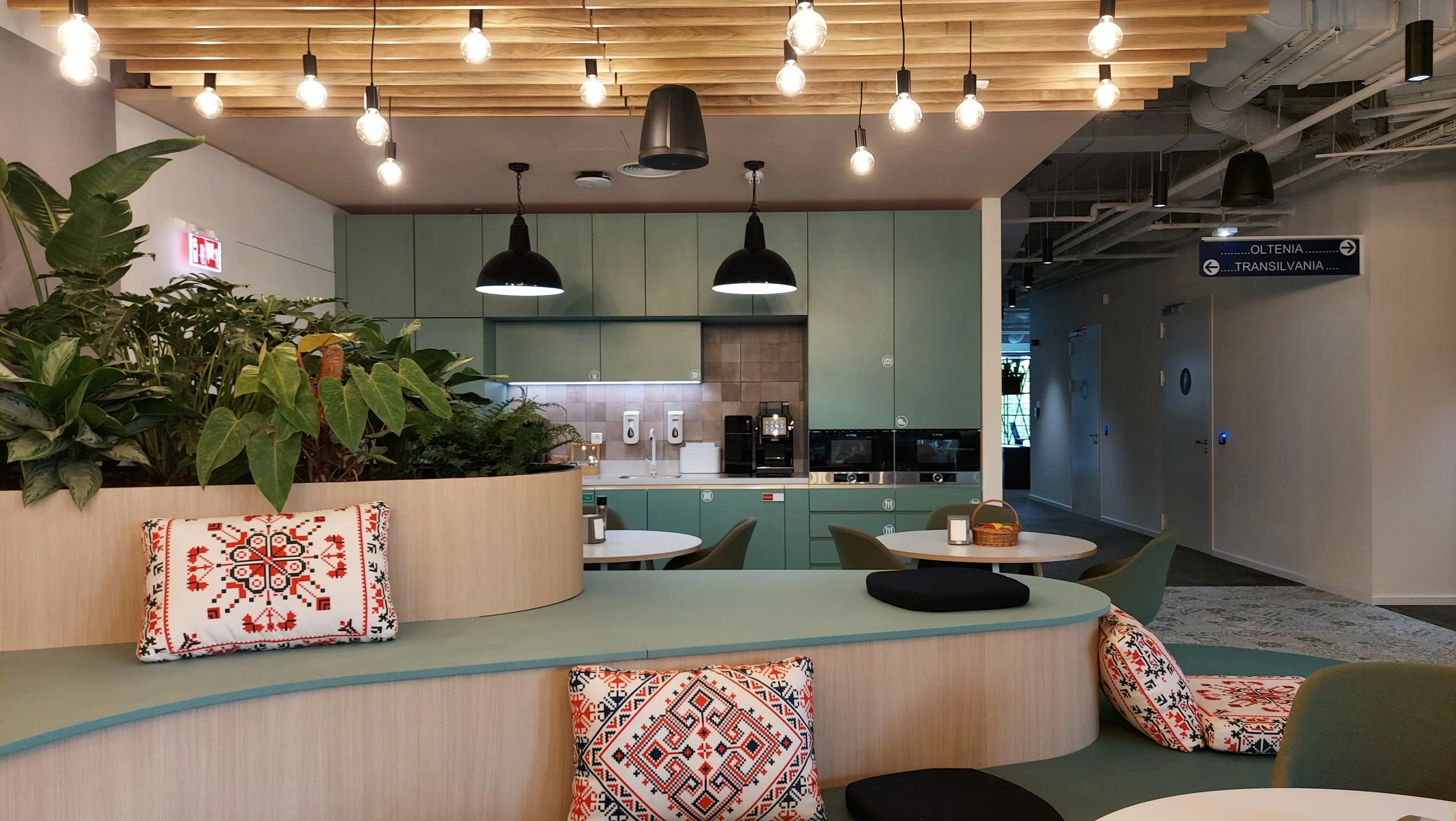 Interior of a modern café with green and wood decor, black pendant lights, green chairs, round tables, and decorative pillows with red and black patterns