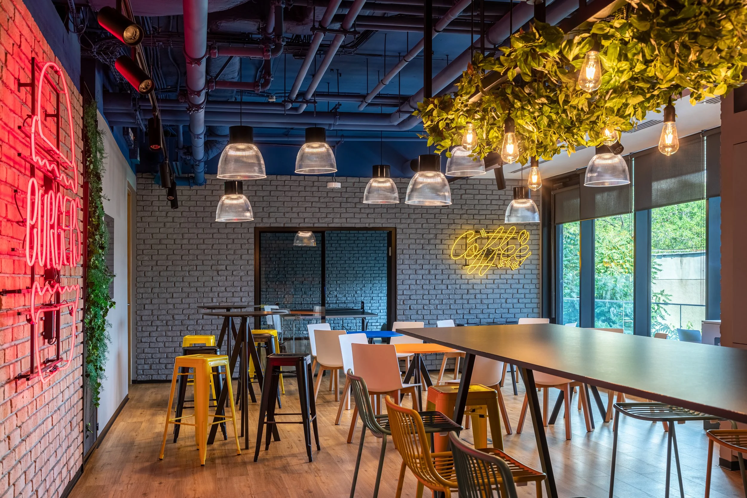 Interior of a modern coffee shop with a mix of high and low tables, colorful chairs, exposed brick walls, and hanging light fixtures. Neon signs reading 'Coffee' and 'Sugar' are displayed, with a large window providing natural light.