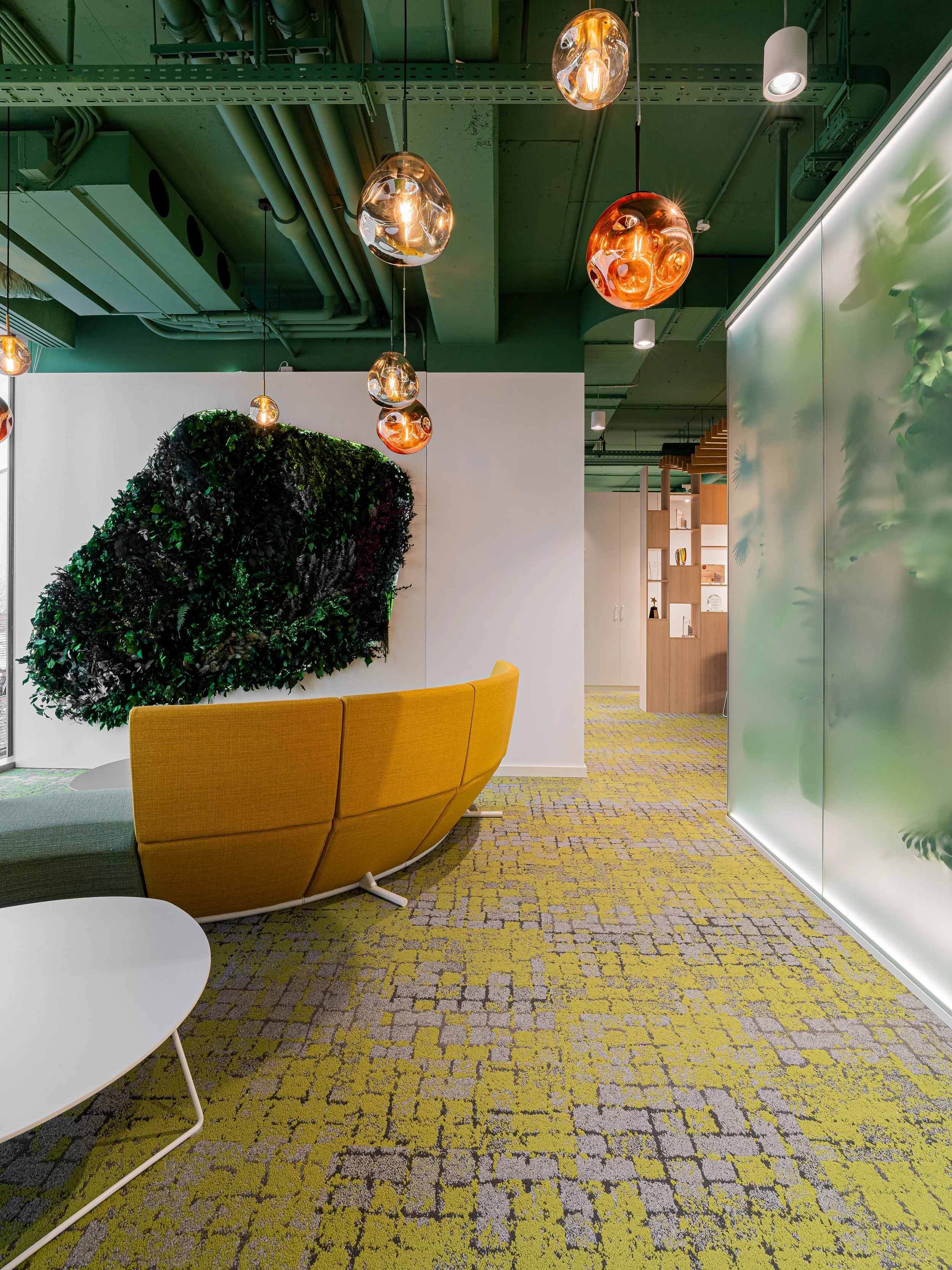 Modern office lounge with yellow curved sofa, white round coffee table, and green moss wall art, illuminated by hanging amber and white pendant lights, and a green ceiling with visible ductwork.