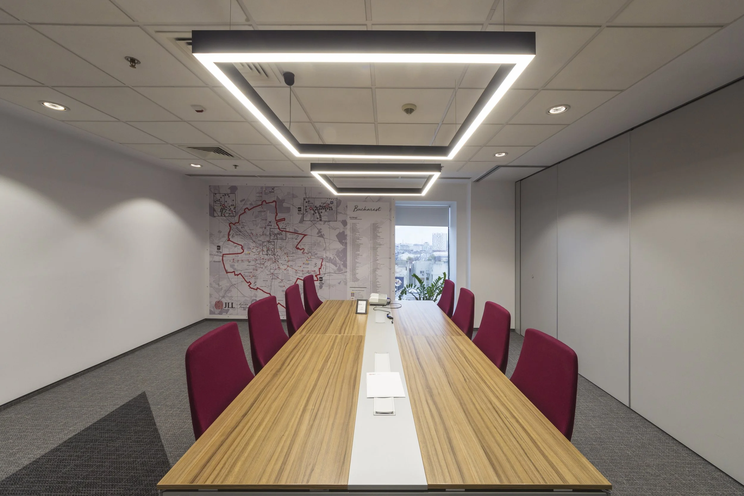 Modern conference room with a large wooden table surrounded by red chairs, a wall with a map of Bucharest, and a window with a city view.