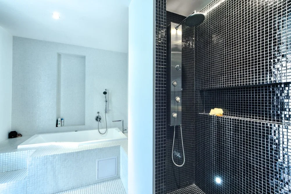 Bathroom with a white bathtub and shower area on the left, featuring white tile walls, and a separate black-tiled shower with a rainfall showerhead and multiple body jets on the right.