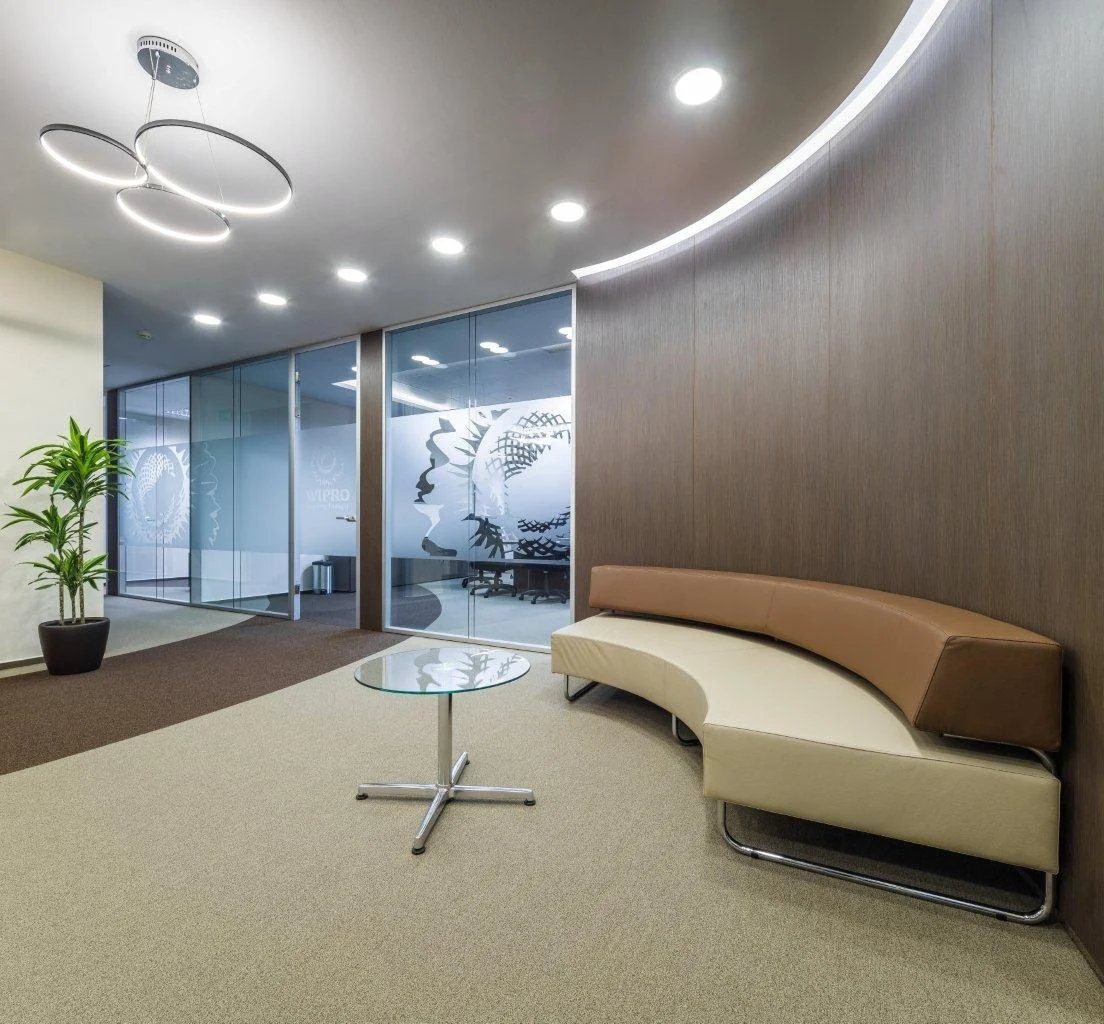 Modern office lobby with ceiling lights, a beige and brown curved sofa, a glass side table, a potted plant, and glass walls with frosted designs.