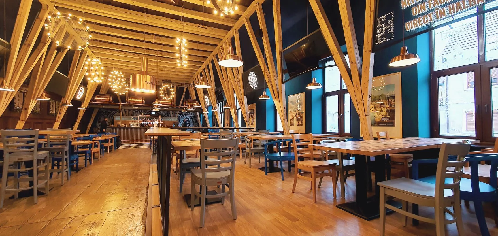 Empty restaurant with wooden tables and chairs, decorated with string lights and framed pictures on blue walls, large windows letting in natural light.