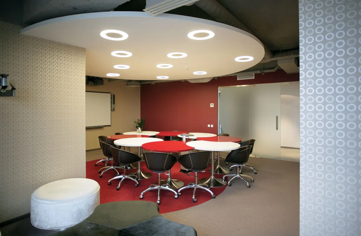 Modern conference room with round red and white tables, black swivel chairs, a white ottoman, a small potted plant, and a whiteboard.