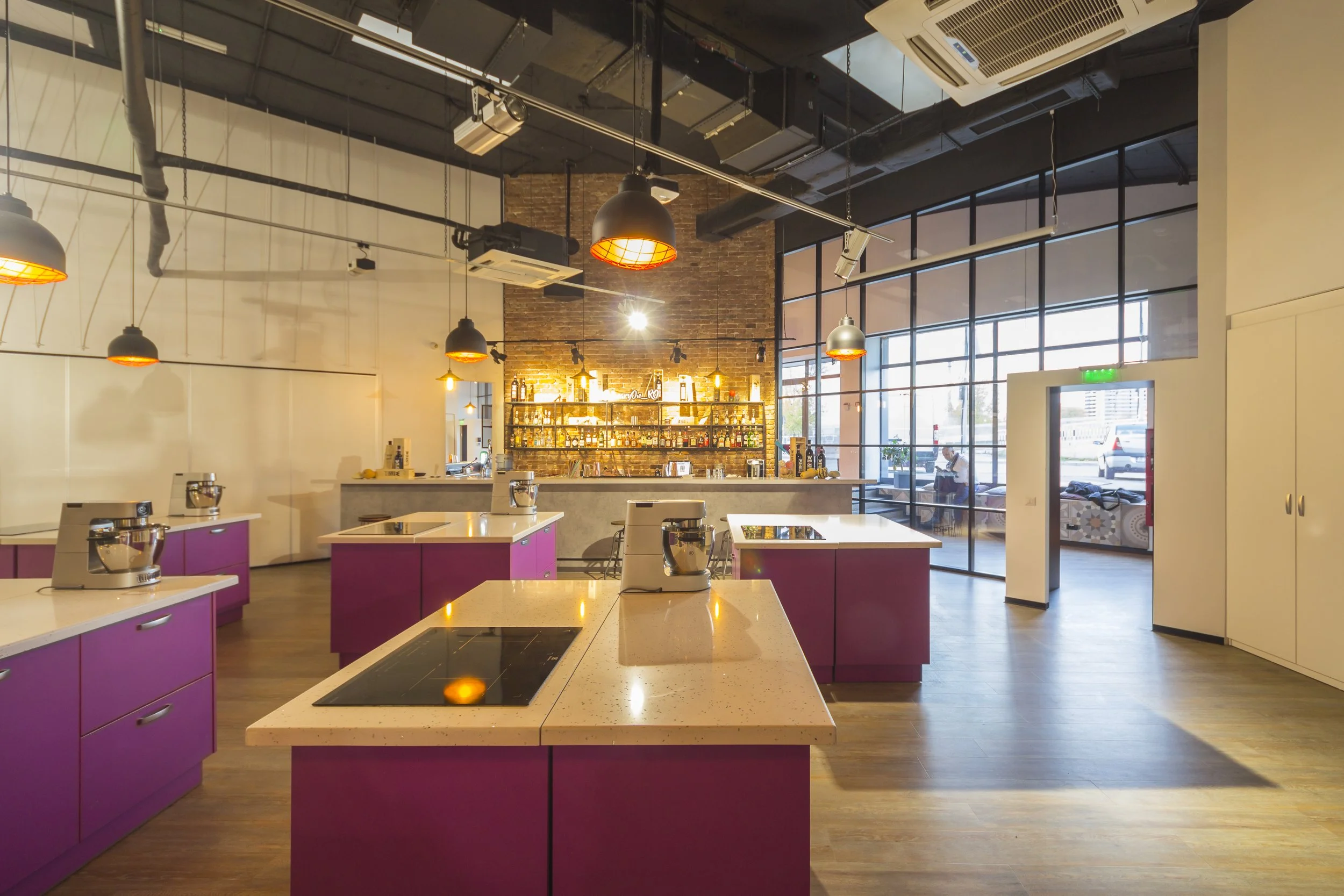 Modern kitchen with purple cabinets, white countertops, and a central stovetop, inside a brightly lit open space with large windows and a lounge area in the background.
