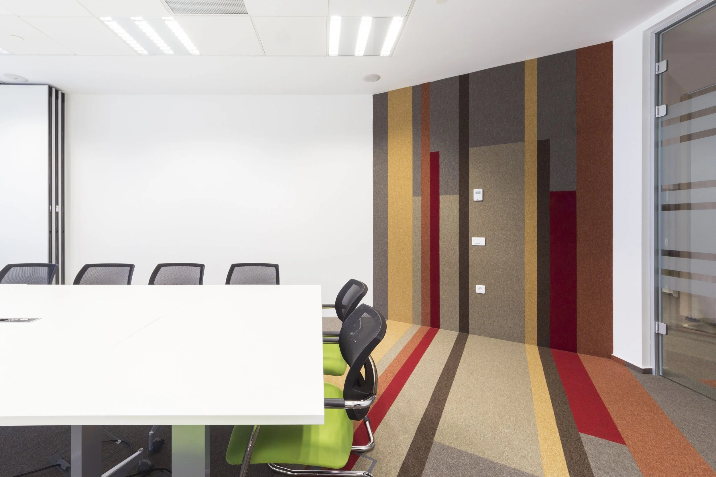 Modern conference room with a white table, black mesh chairs, green chair accents, and a colorful striped carpeted wall with electrical outlets.