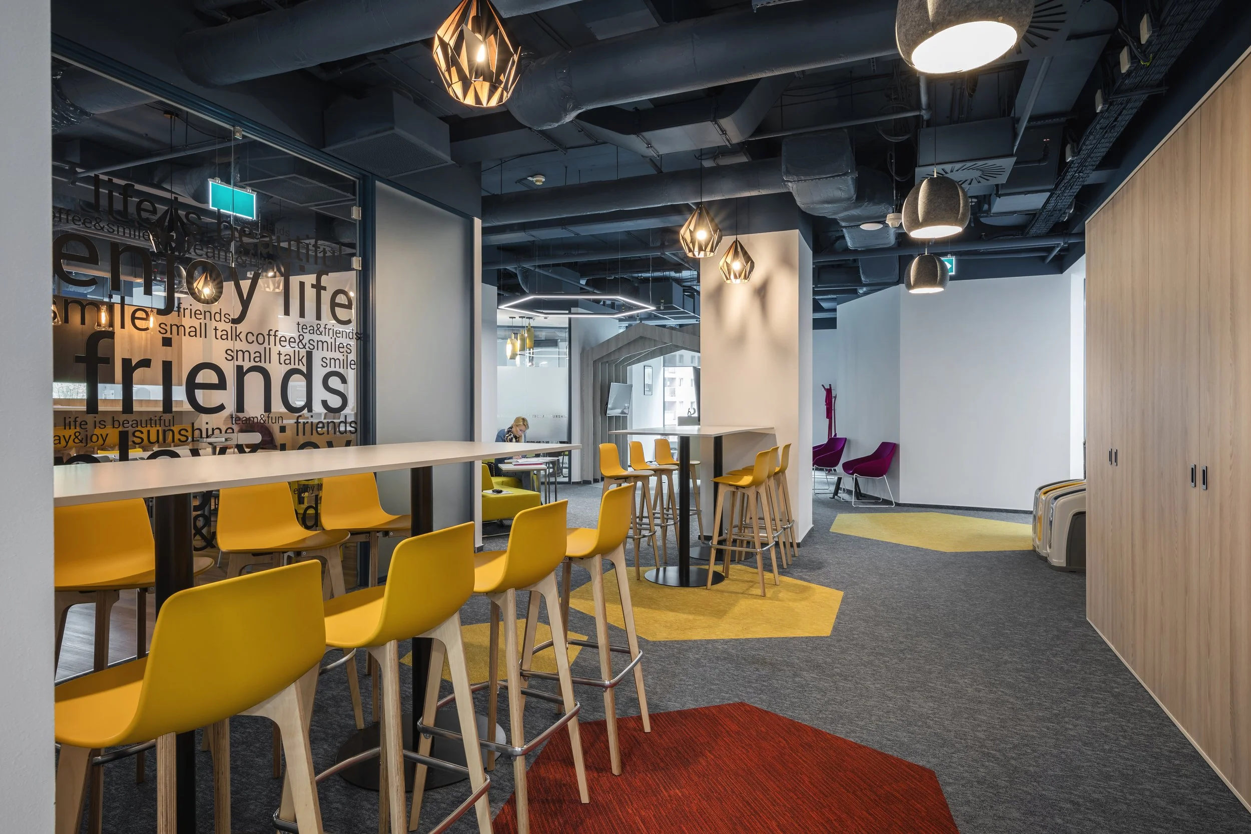 Modern office break area with yellow bar stools around a high table, colorful carpet patches, and a seating area with chairs in purple and yellow, illuminated by hanging pendant lights in a contemporary style.