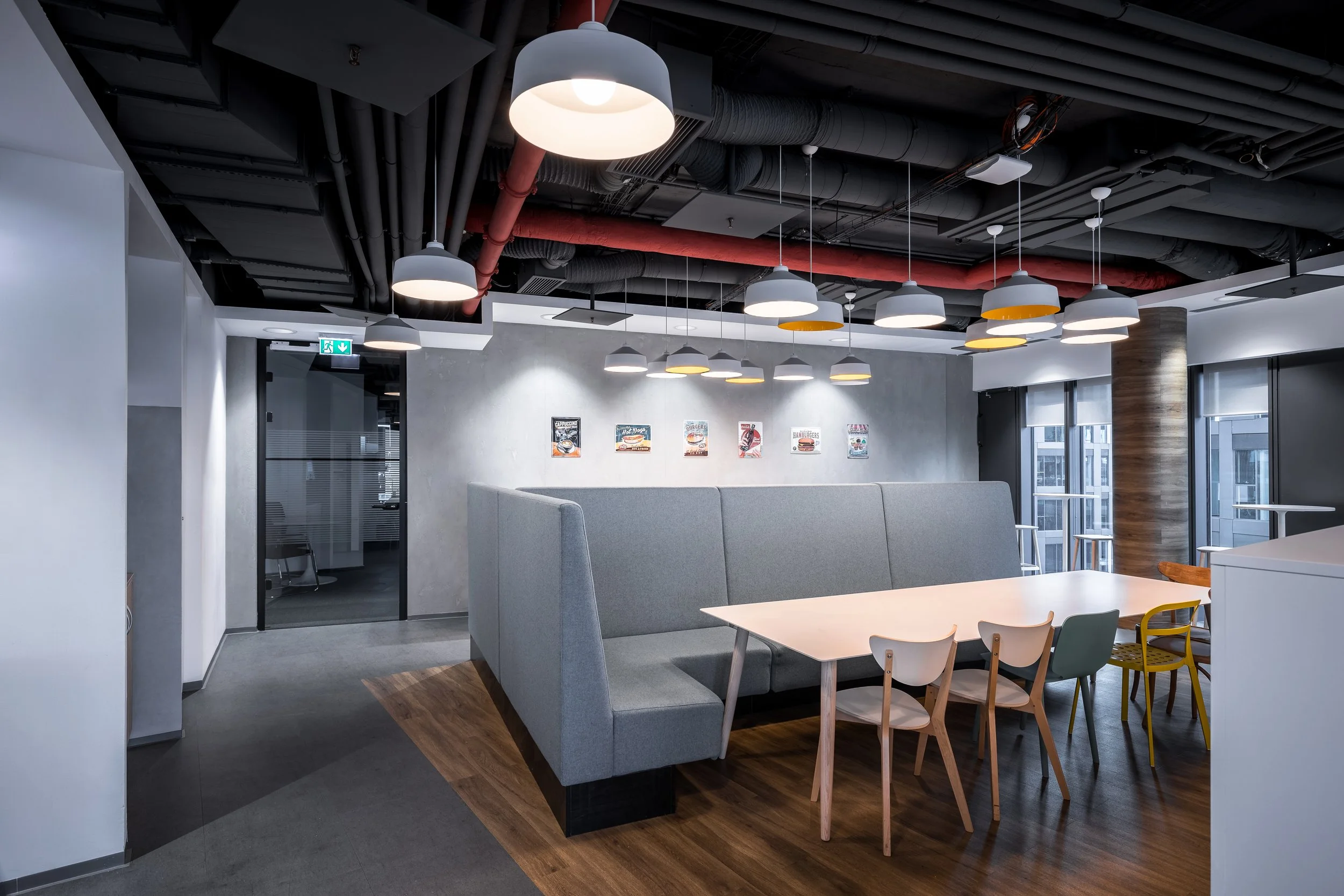 Modern office break room with a large white table, assorted chairs, gray high-back booth seating, contemporary pendant lighting, a textured wall with posters, and large windows revealing city buildings.