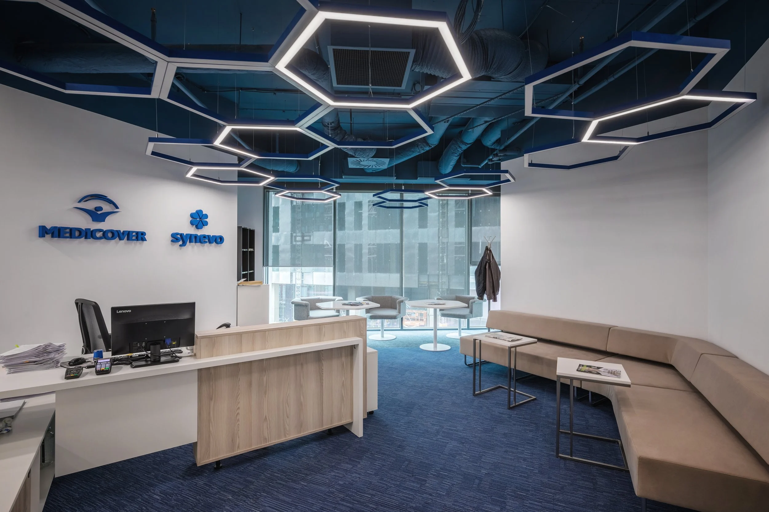 Office reception area with a white desk, a computer, and office supplies on it. The wall displays the logos of Medicover and Synevo in blue. There is a beige curved sofa, small side tables with magazines, and a seating area with windows in the background. The ceiling features modern hexagonal light fixtures.