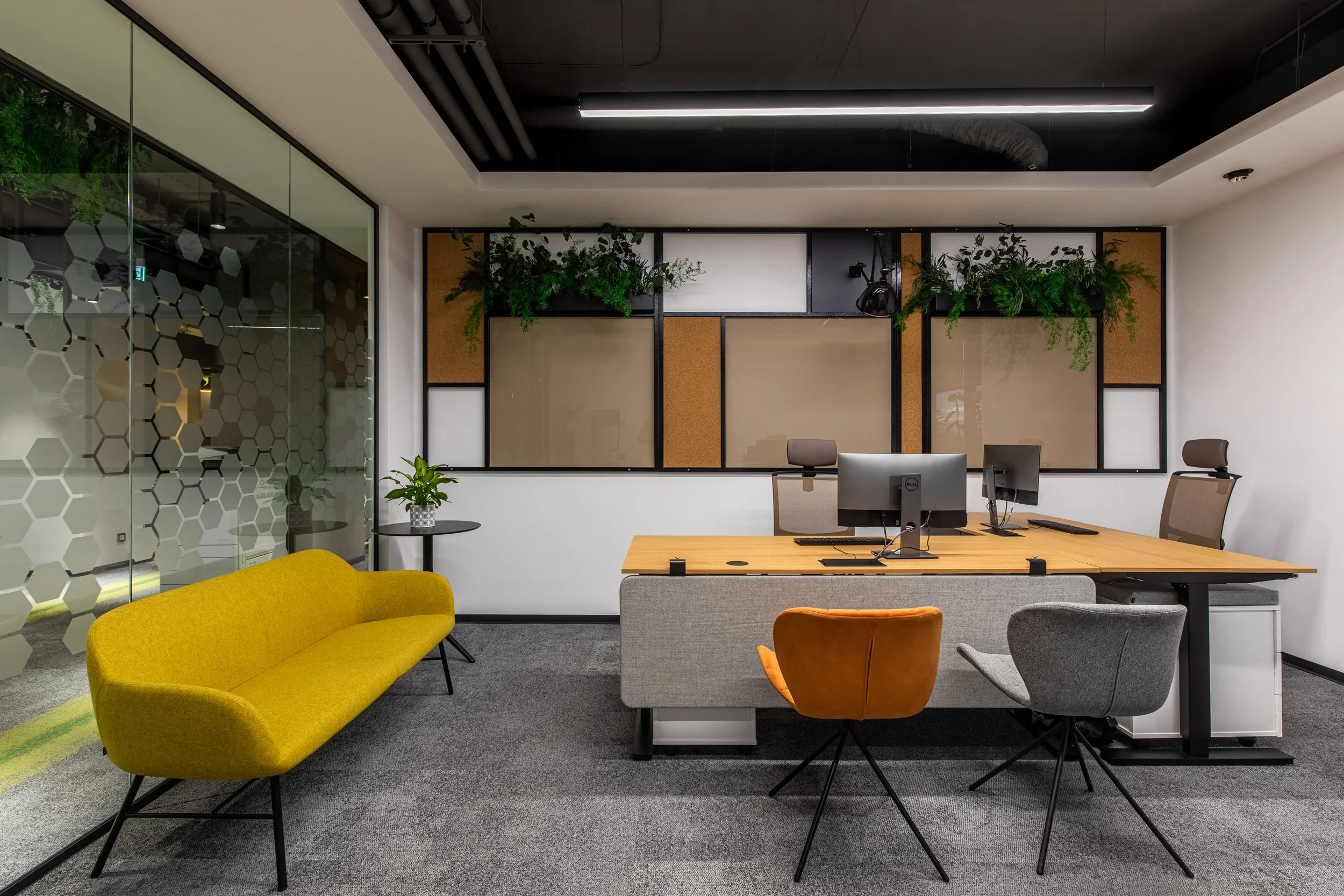 Modern office space with a yellow sofa, a wooden desk with computer monitors, and several chairs, including gray and orange ones. There are plants on a wall with a wooden and cork design and a glass wall with a hexagon pattern.