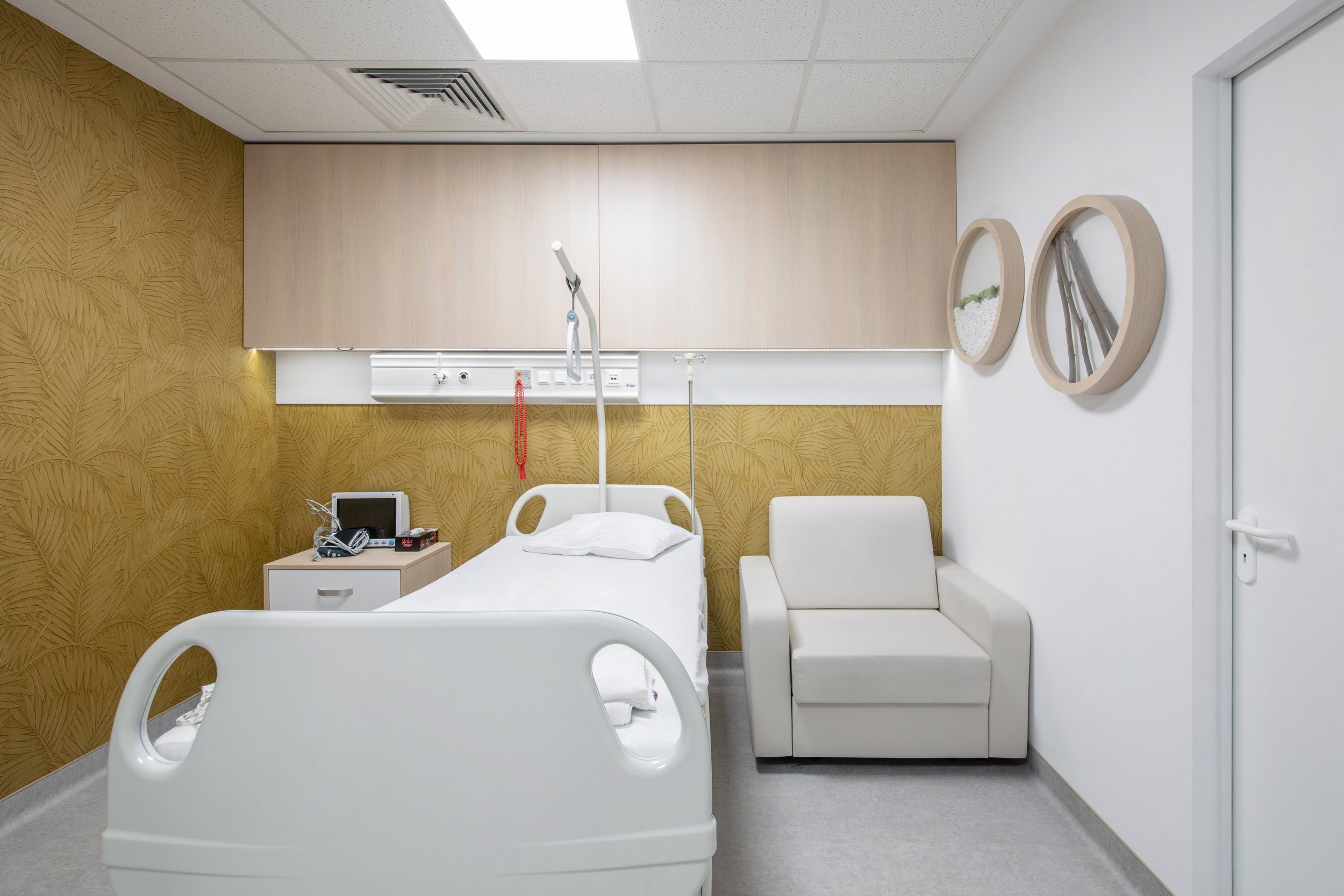 A hospital room with a hospital bed, a side table with medical equipment, a white armchair, and decorative wall art. The bed has guardrails and medical devices attached, with a pillow on the mattress.