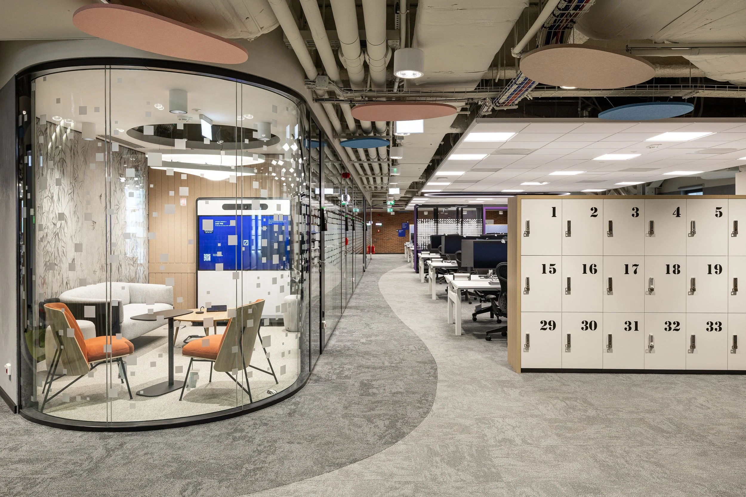 Modern office space with a glass-walled meeting room, office desks, and lockers.