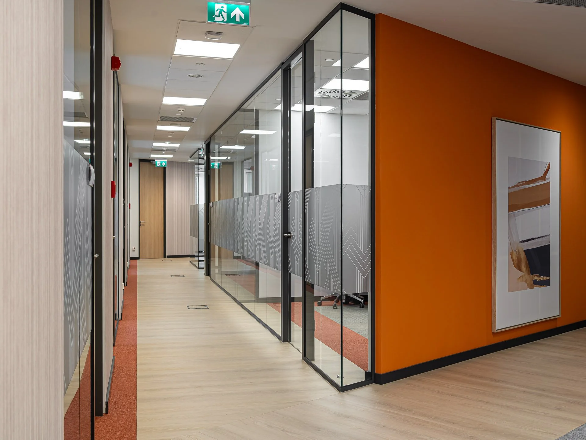 Office corridor with glass-walled meeting rooms, orange wall, artwork, and ceiling exit sign.