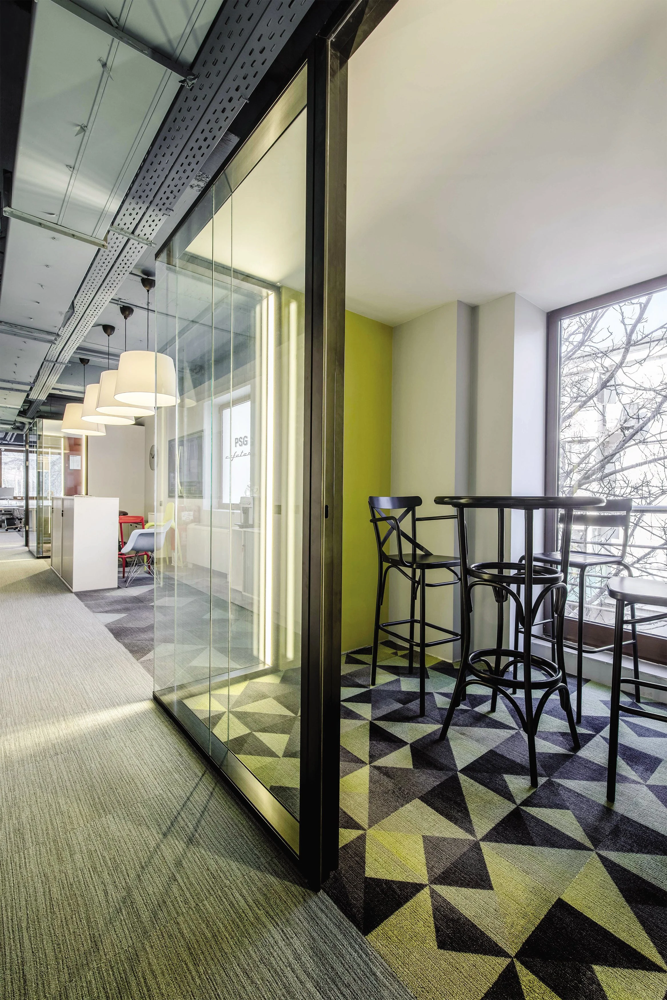 Office space with a glass wall, black high chairs, and a window with trees outside. Modern decor with geometric patterned carpet.