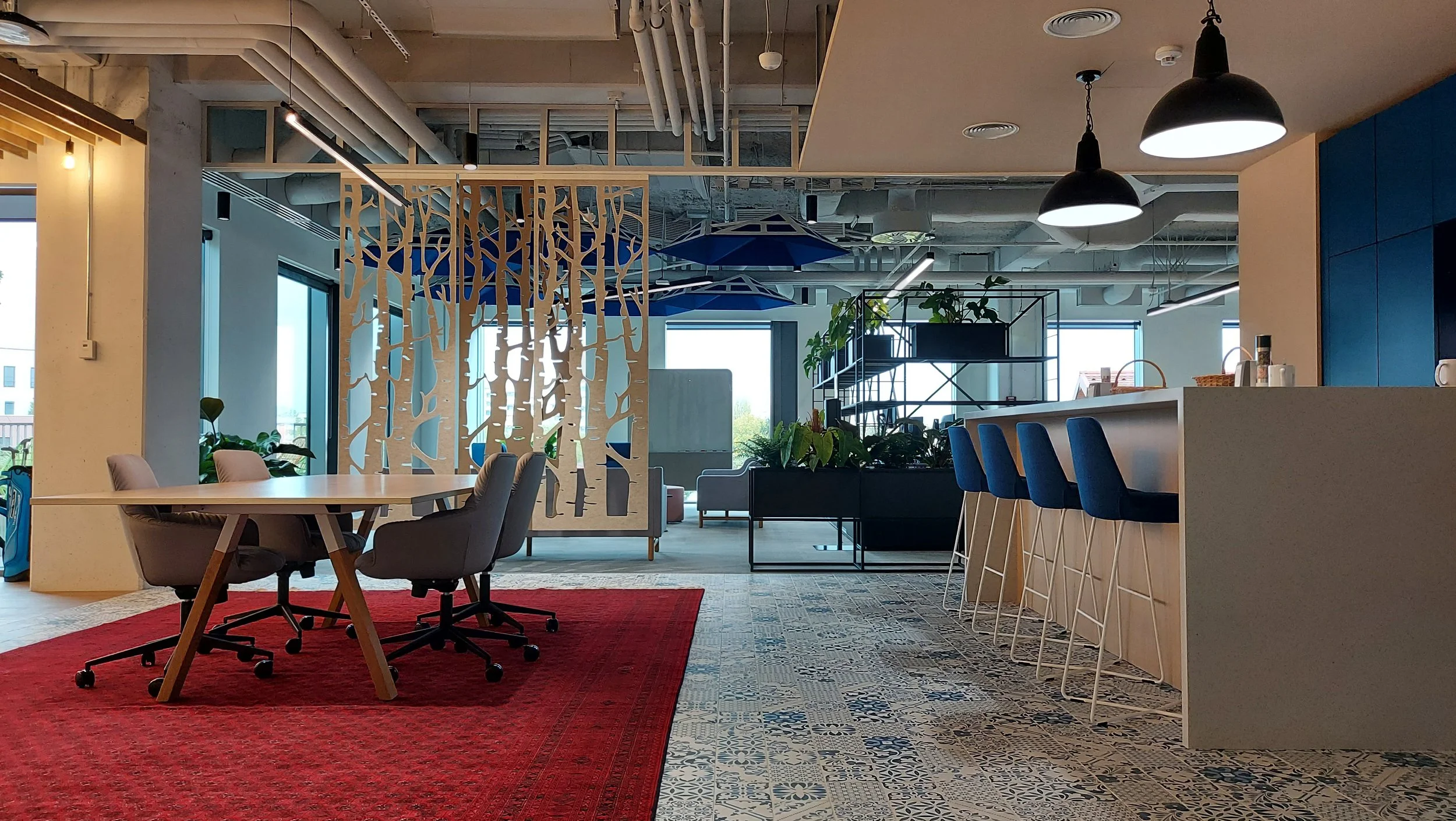 Modern office space with a meeting area, bar-height counter with blue chairs, a table with office chairs, patterned tile flooring, and decorative room dividers.