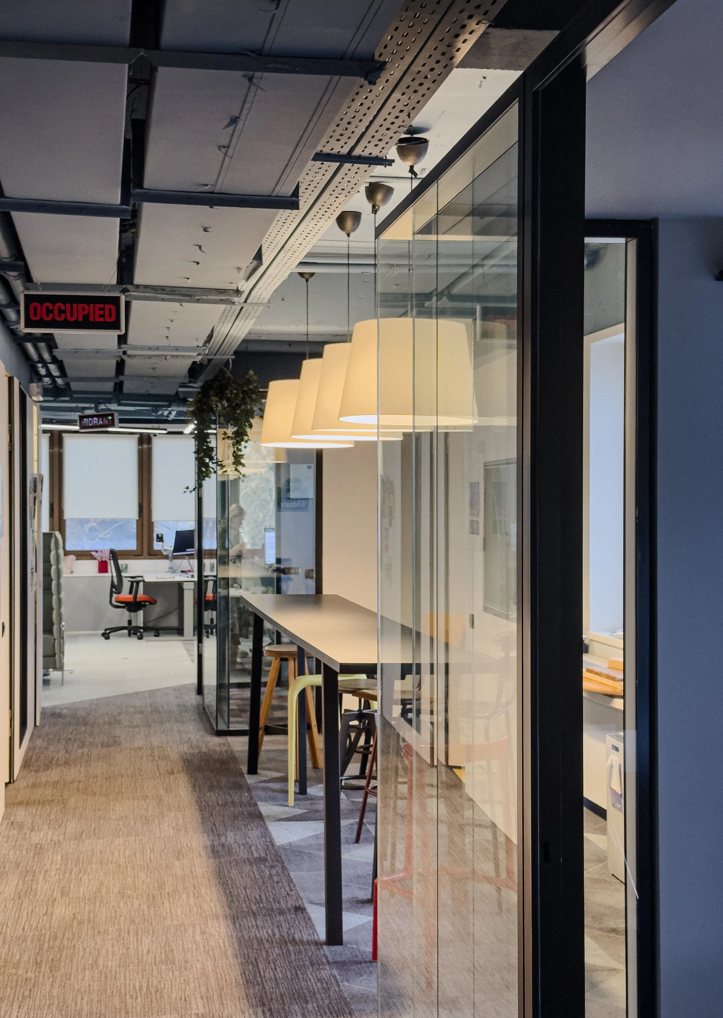 A modern office hallway with a glass partition, hanging ceiling lights, and various chairs and desks visible through the glass.