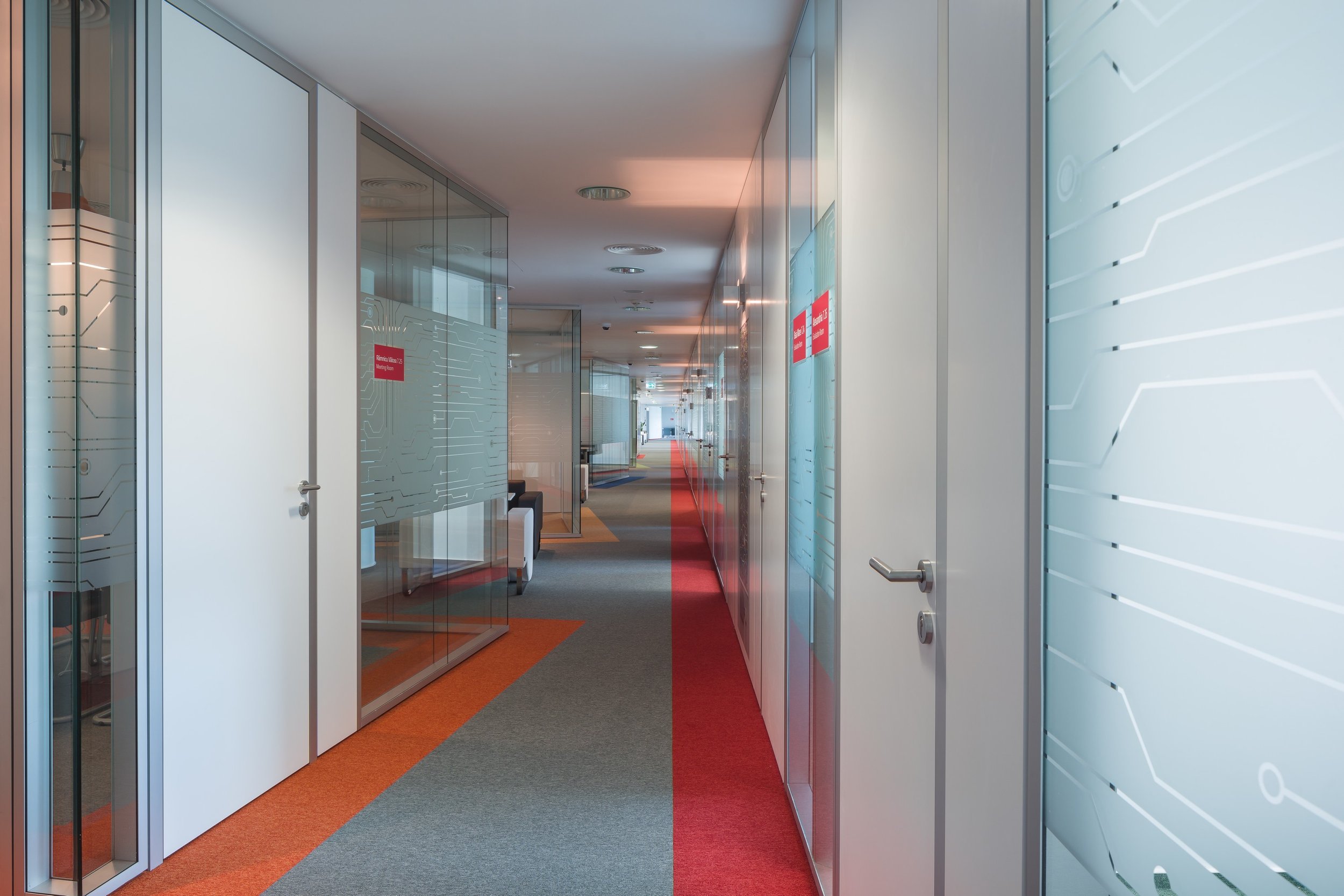 Office corridor with glass-walled rooms, gray, orange, and red carpet, modern furniture, and digital graphics on the walls.
