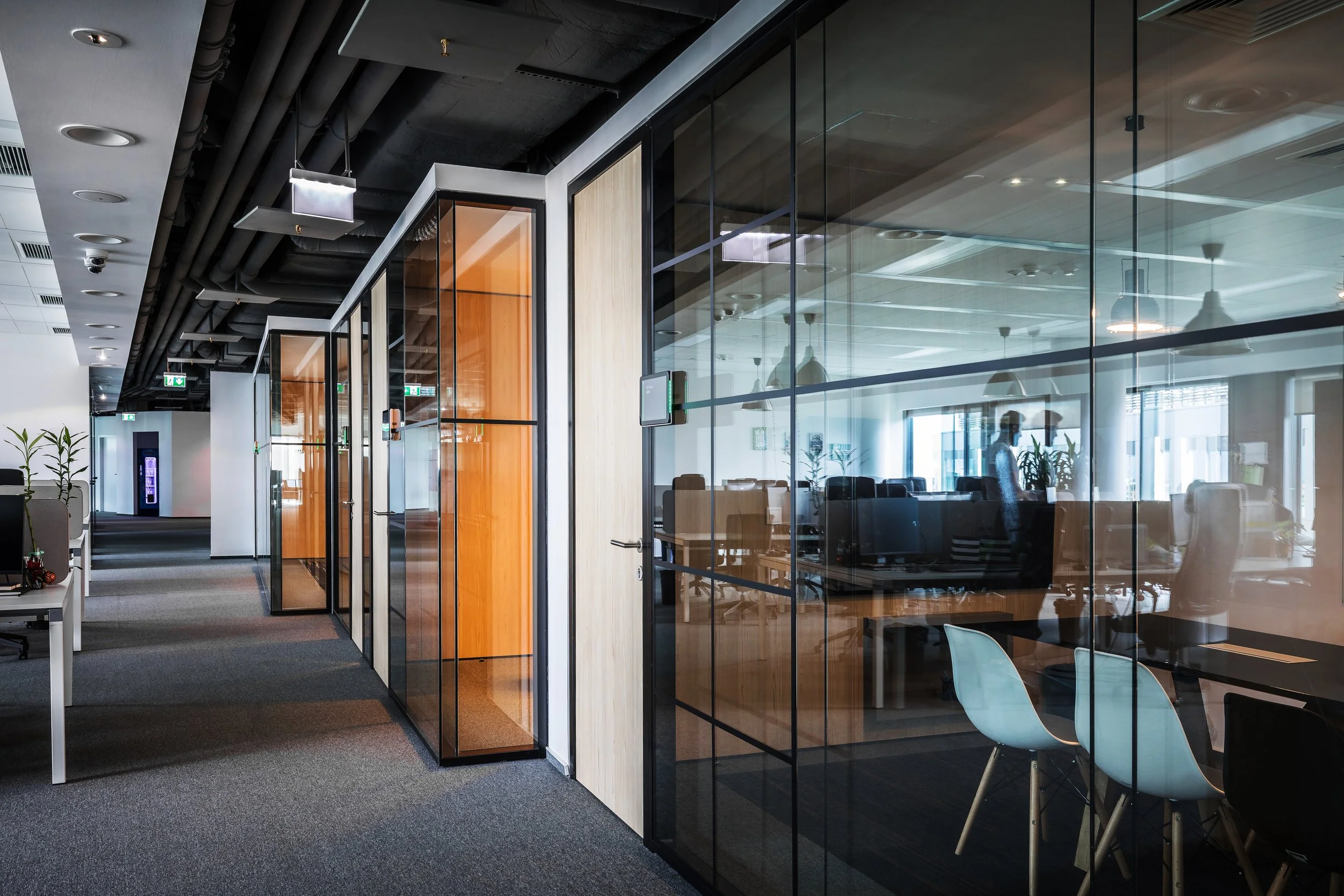 Modern office corridor with glass office rooms, black ceiling with exposed ductwork, and gray carpeted flooring.