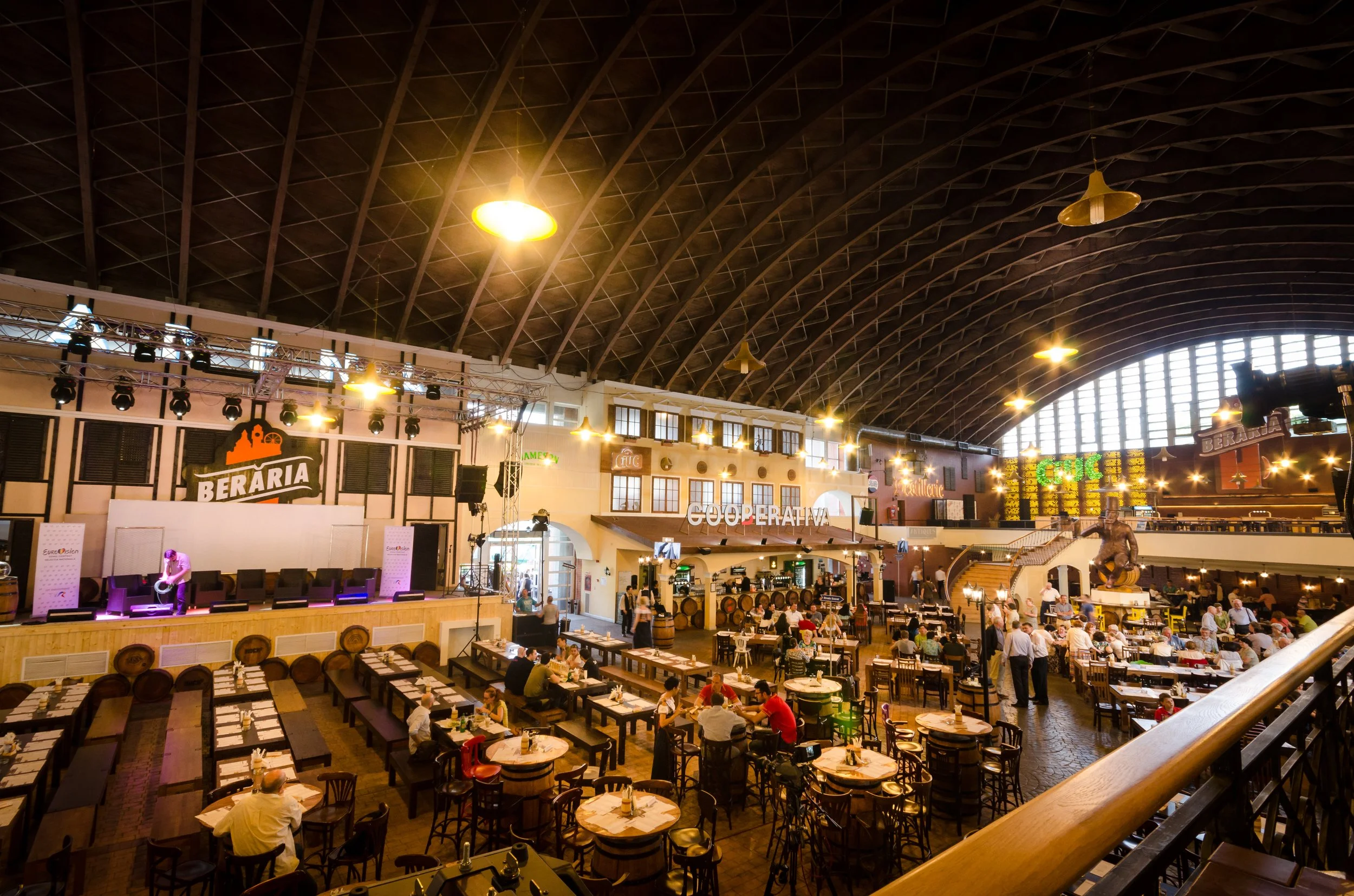 Interior of a large restaurant with a stage on the left, tables and chairs filling the space, people dining, and bright overhead lighting in a spacious hall with high arched ceilings.