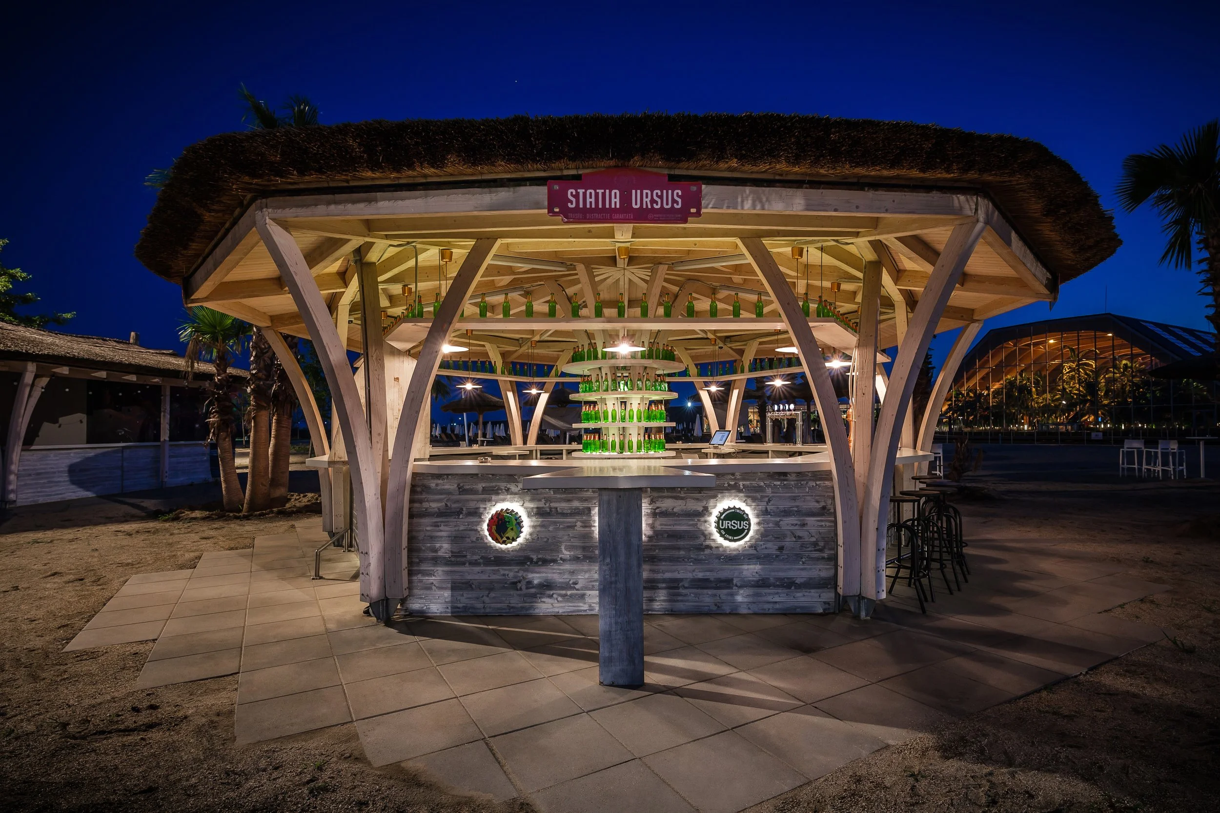 Nighttime view of a wooden bar structure with a thatched roof, decorated with green bottles, and illuminated by lights, with a sign reading 'Statia Ursus' at the top.
