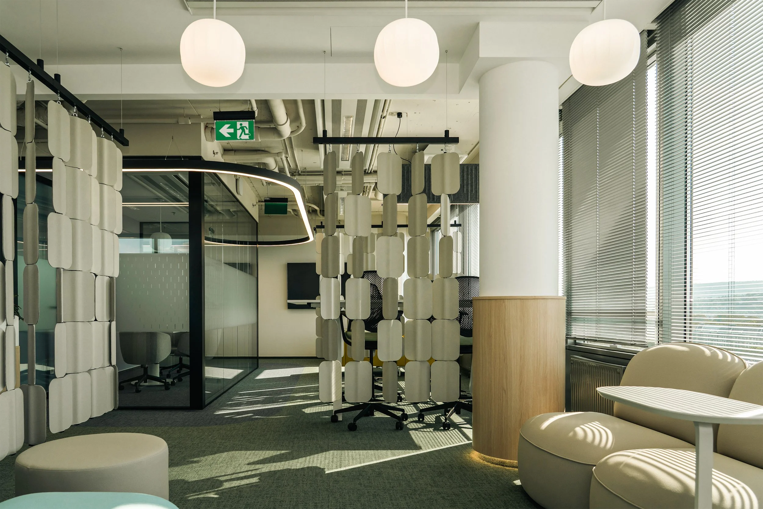 Office space with large windows and blinds, beige seating with a small table, decorative dividers, and chairs in a glass-enclosed meeting room
