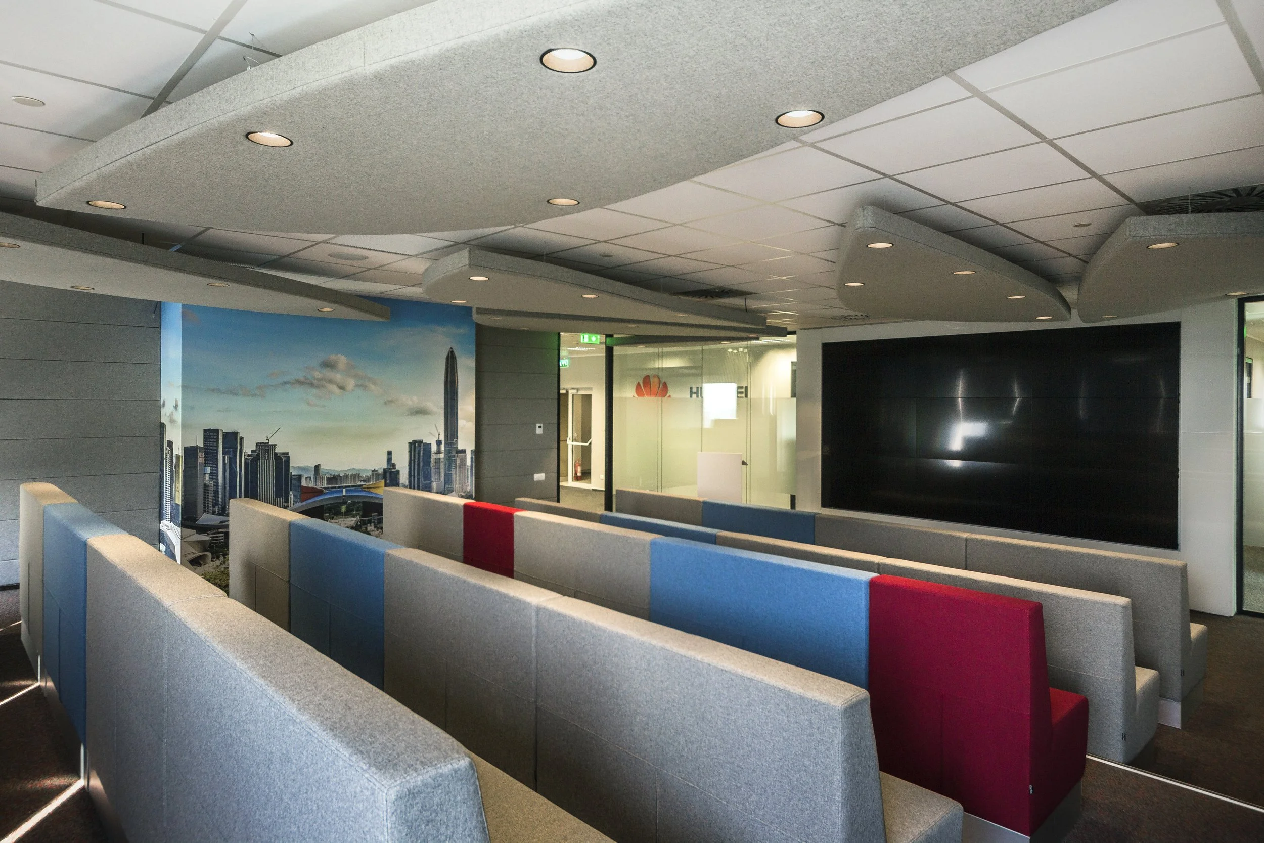 Empty modern office lounge with colorful seating, a large black television, and a cityscape mural, near a glass door with Huawei logo.