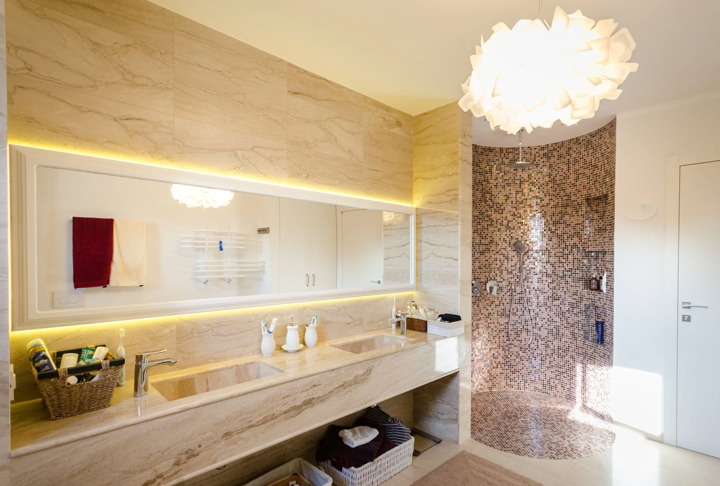 Bathroom with dual sinks, large mirror, beige marble surfaces, and a walk-in shower with brown mosaic tiles.