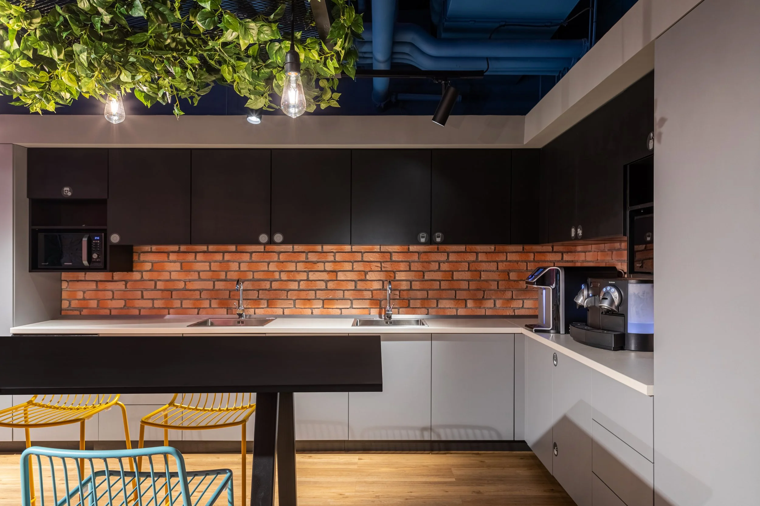 Modern kitchen with black upper cabinets, gray lower cabinets, brick backsplash, two sinks, a coffee station, microwave, and lightbulb hanging from ceiling with green leaves above.