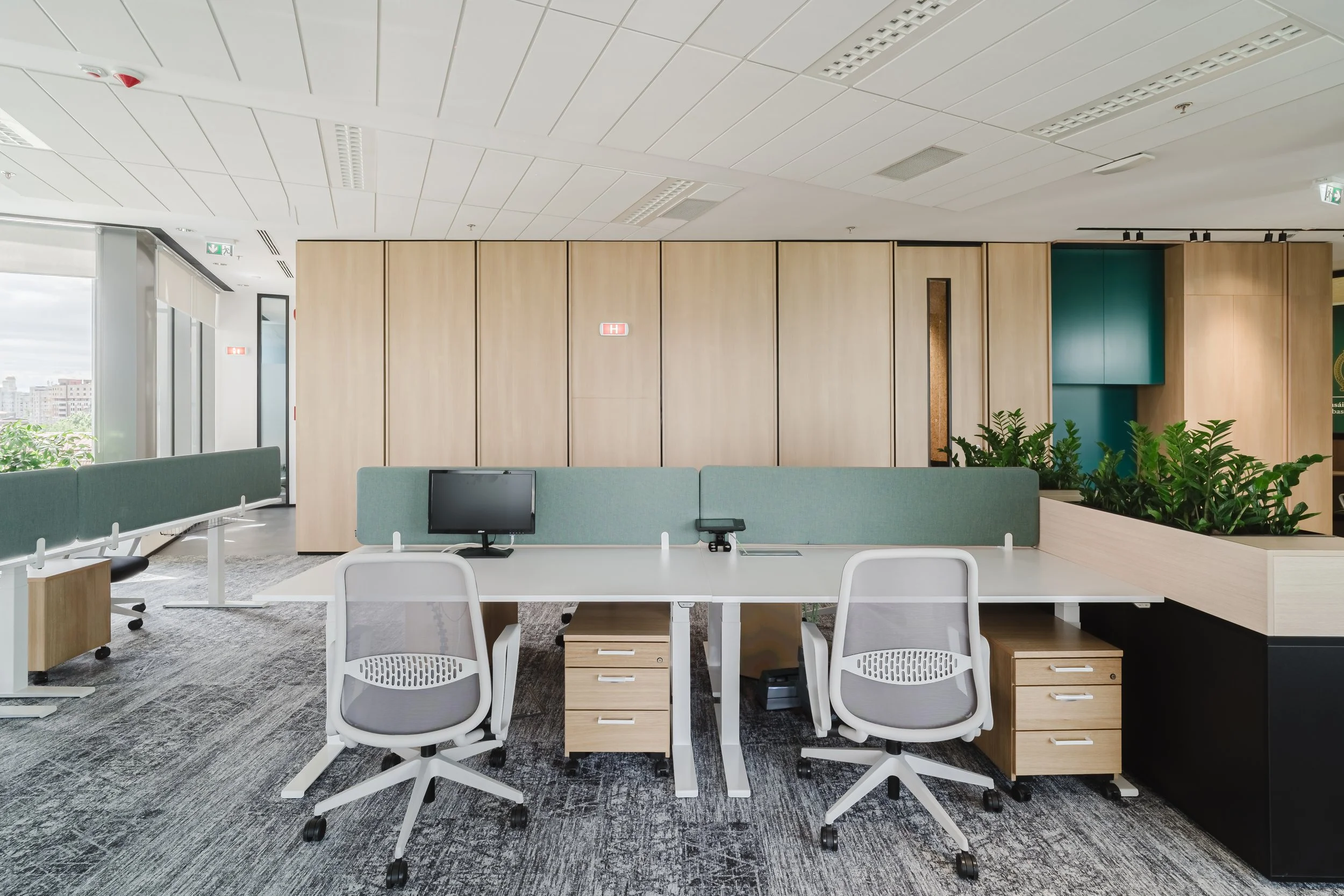 Modern office workspace with two white swivel chairs, a desk with a partition, a computer monitor, and small wooden drawers, surrounded by planters with green plants, large windows with cityscape view, and wooden wall panels.