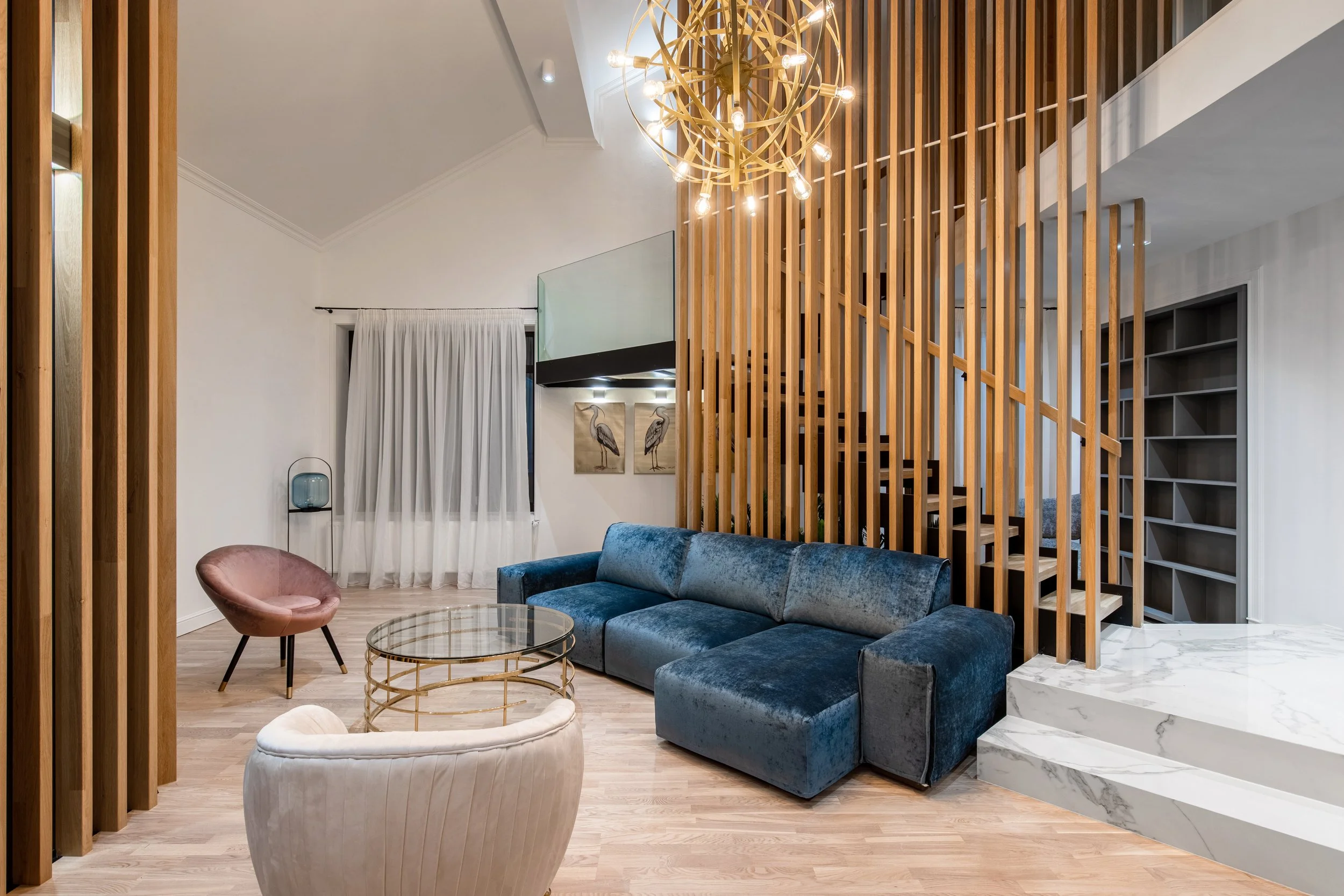 Modern living room with a navy blue velvet sectional sofa, pink accent chair, glass-top coffee table with gold frame, white upholstered armchair, wooden slat wall, white curtains, and a spherical gold chandelier.