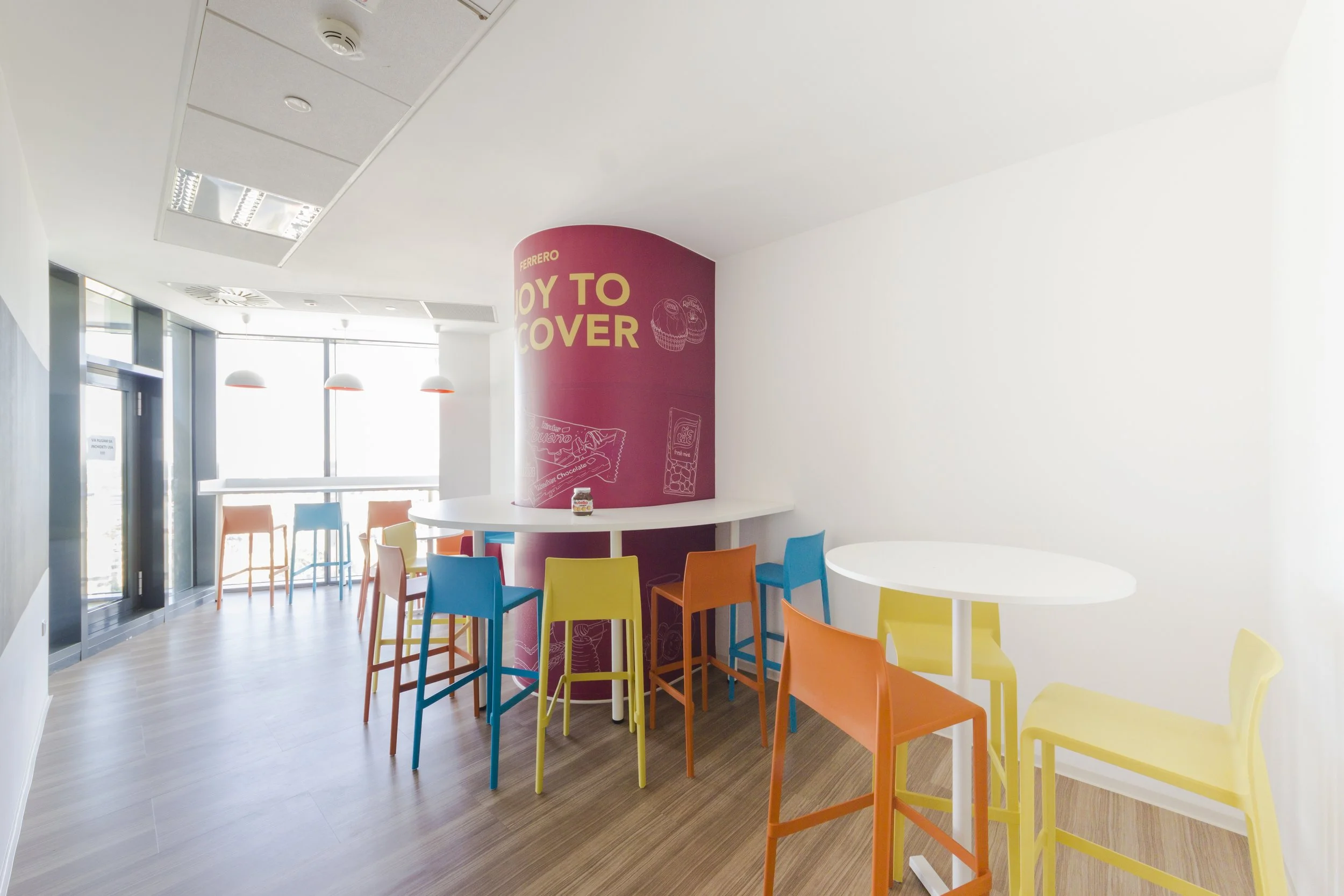 Bright, modern cafe interior with colorful chairs and white tables near large windows, with a pink cylindrical promotional display in the center.