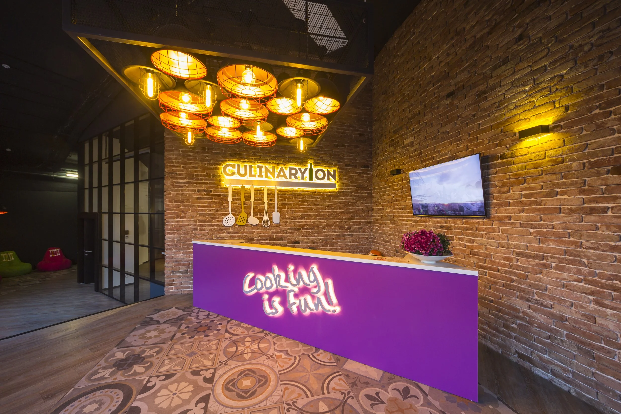 A modern restaurant or kitchen-themed reception area with exposed brick walls, colorful floor tiles, and vibrant lighting. There is a purple reception desk with a neon sign that says 'cooking is fun!', a flower arrangement, and a flat-screen TV on the wall. Above, there is a decorative lighting fixture with warm-toned bulbs, and a neon sign that reads 'CULINARY ON' with utensils hanging underneath.