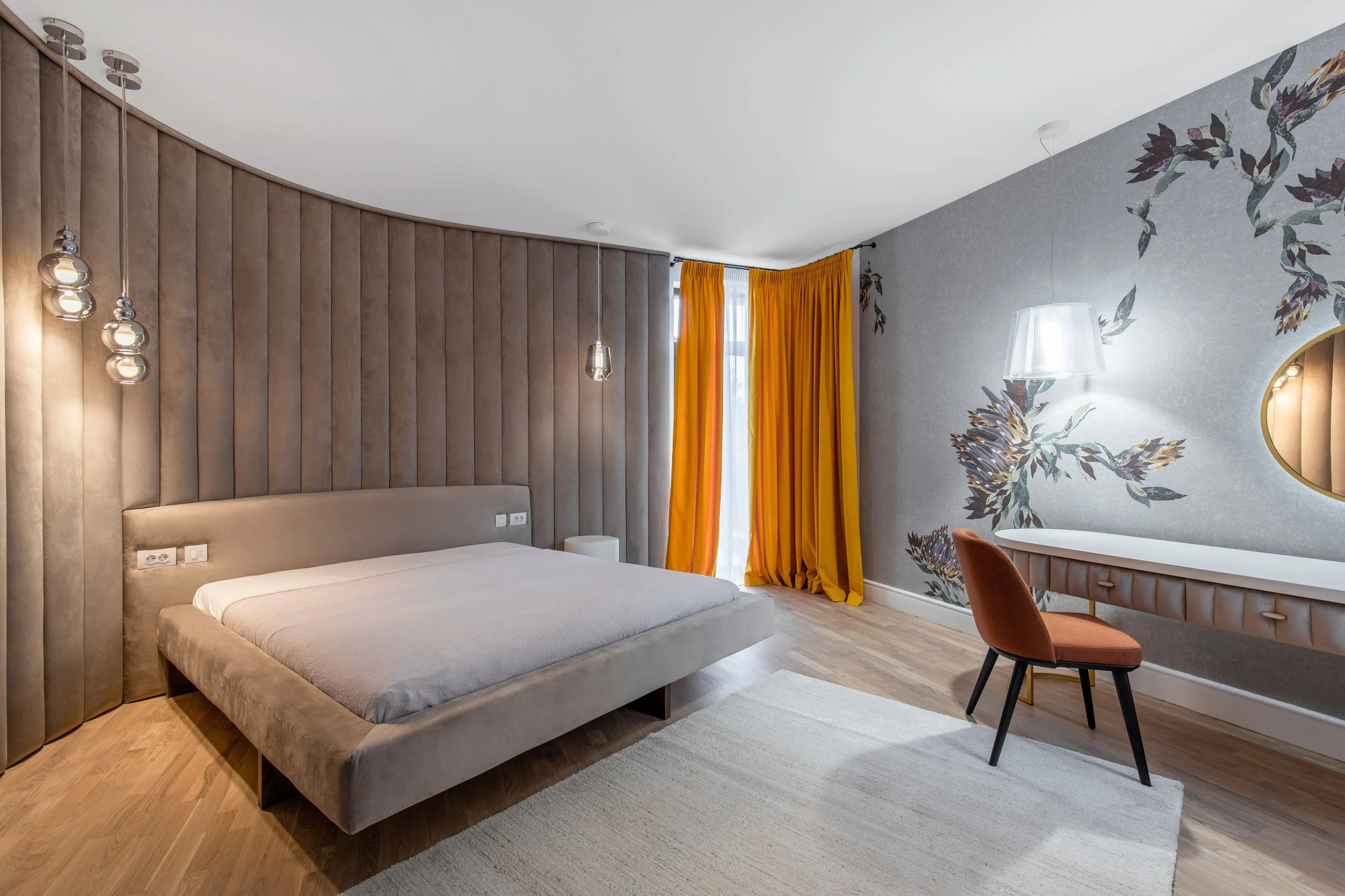 Modern hotel bedroom with a large bed, yellow curtains, and a floral accent wall. Decor includes hanging pendant lights and a small desk with a chair.