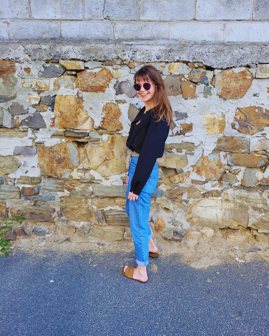 A woman with brown hair wearing sunglasses, a black top, a black blazer, blue jeans, and brown sandals standing against a weathered stone wall, smiling.