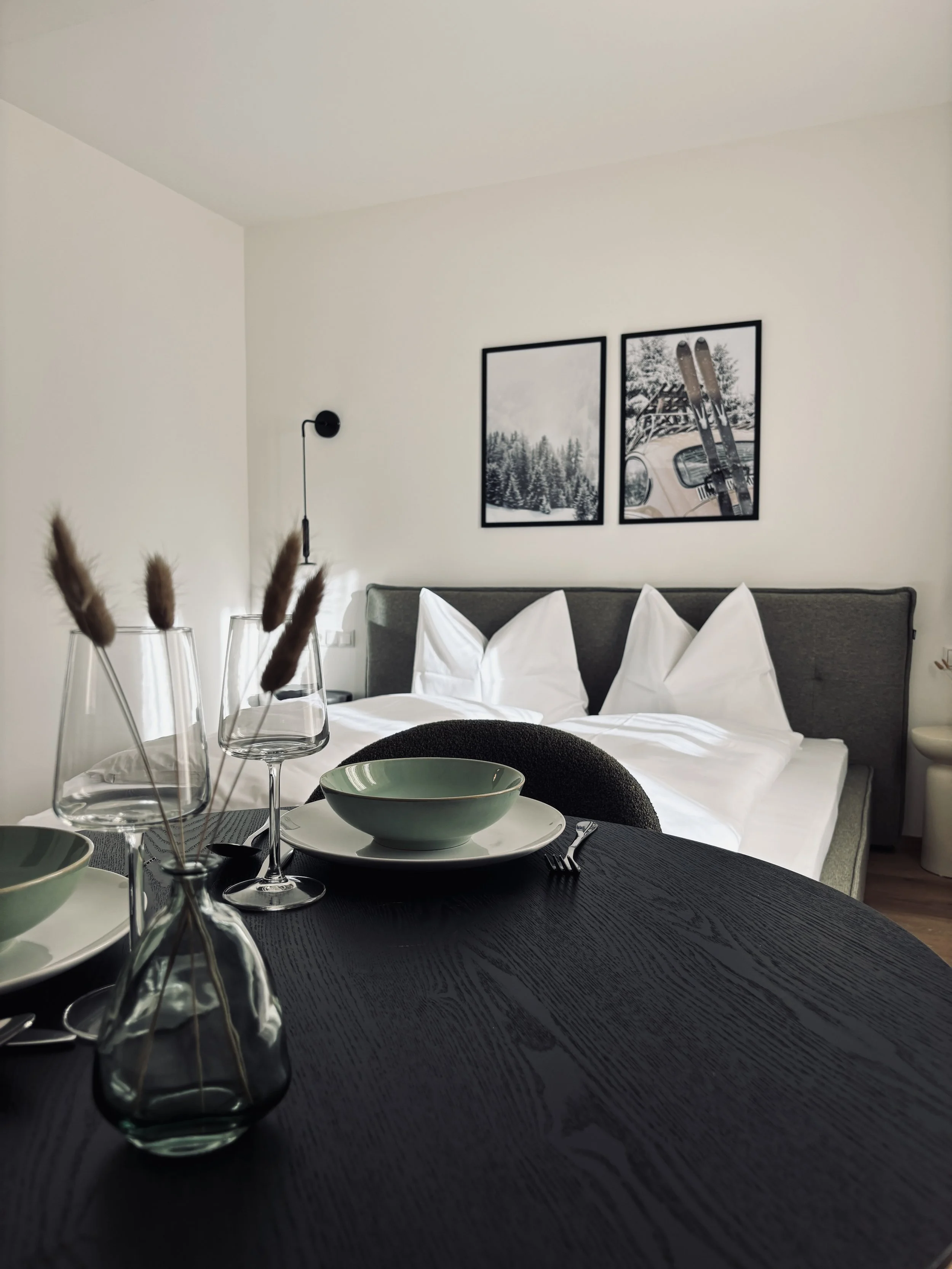 Modernes Schlafzimmer mit doppeltem Bett, weißen Kissen, Wandbildern mit Schneelandschaft und Skiern, Esstisch mit Geschirr und Dekoration, dunklem Holz Tisch.