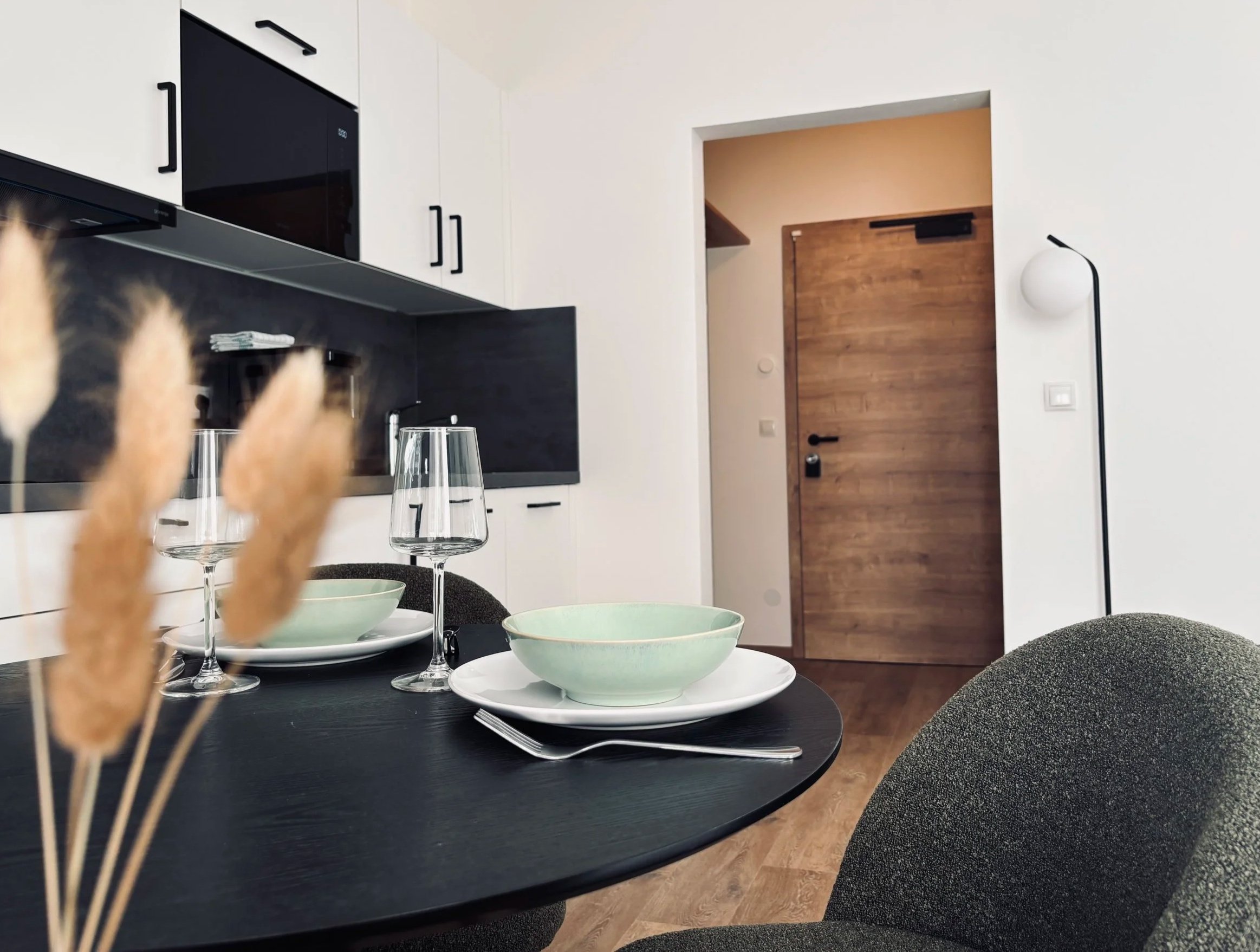 Ein moderner, minimalistisch eingerichteter Essbereich mit schwarzem Tisch, grünen Schalen, Wein glasses, dunklen Stühlen und einem weißen Wand mit Holz Tür im Hintergrund.