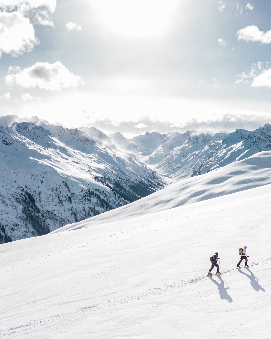 Zwei Skifahrer auf schneebedehnten Bergen mit hohen, schneebedeckten Gipfeln im Hintergrund, sonniger Himmel und einige Wolken.